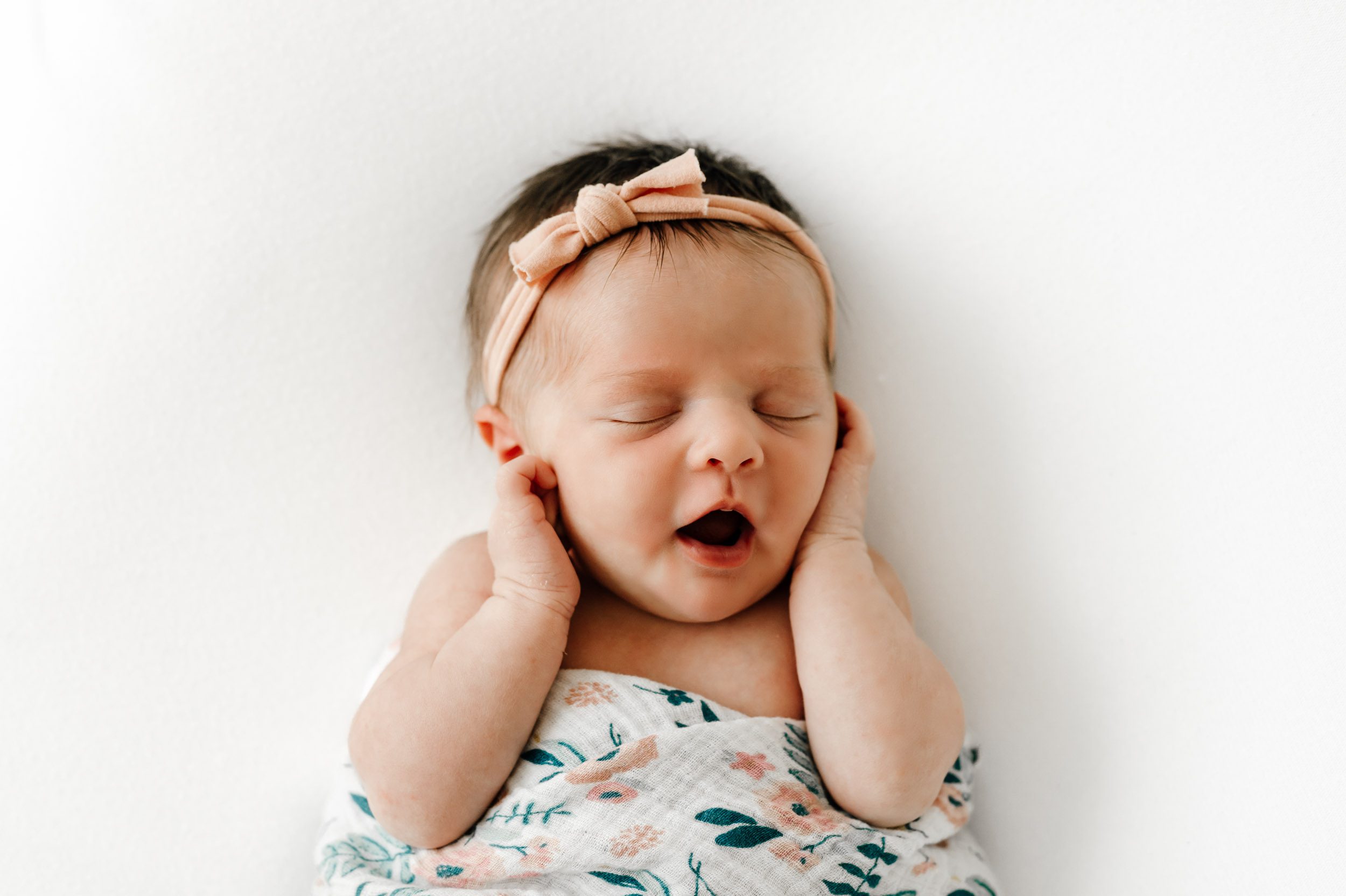 a newborn baby girl laying on her back on a white backdrop holding her hands up to her face with her mouth open wide during a studio newborn photography session