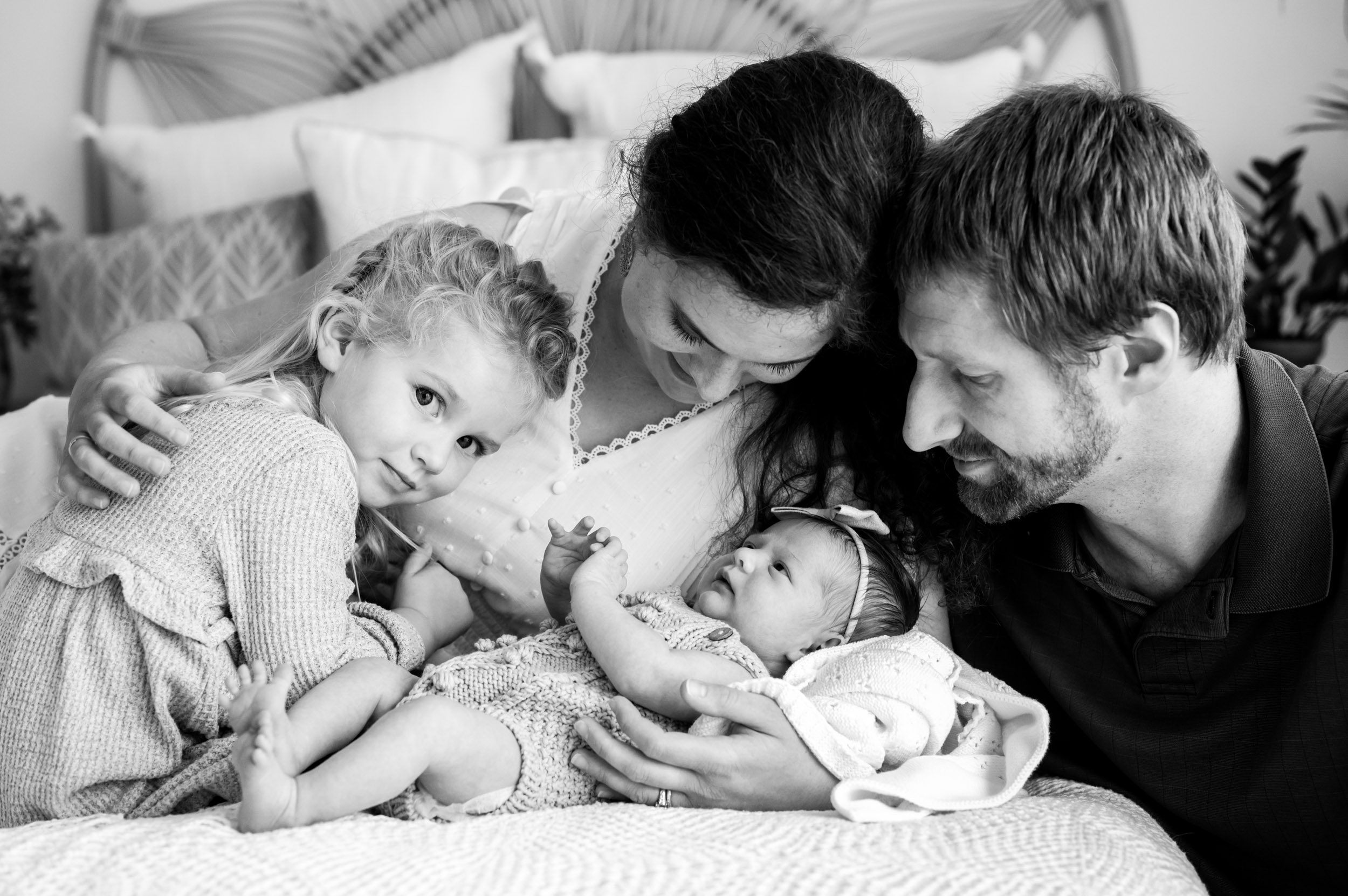 a black and white picture of a new family of four laying on a bed with their baby girl while mom and dad rest their foreheads together and their older daughter looks right at the camera with a hint of a smile on her face during a Pottstown studio newborn photoshoot