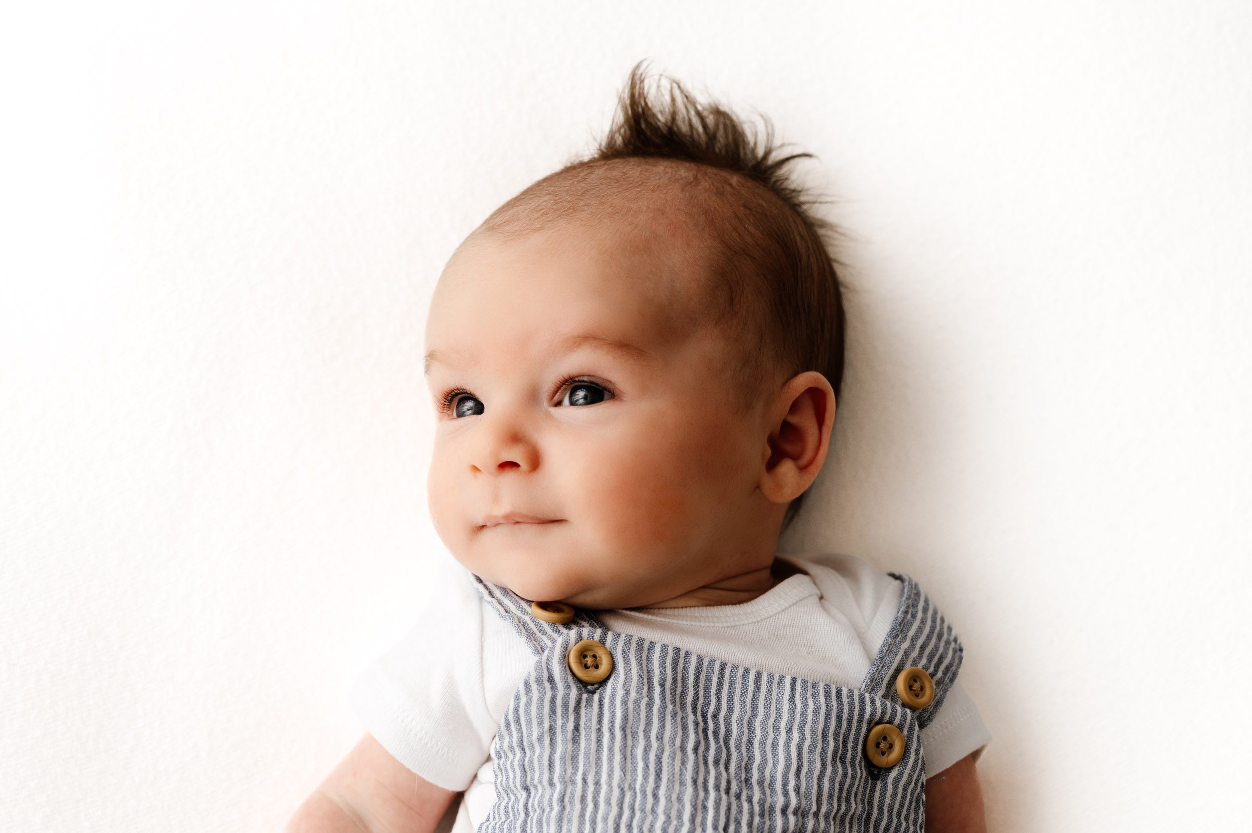 an 8 week old baby boy wearing a blue and white striped romper laying on a white backdrop and looking off toward the window light on the left to show off his baby mohawk during a newborn photos session