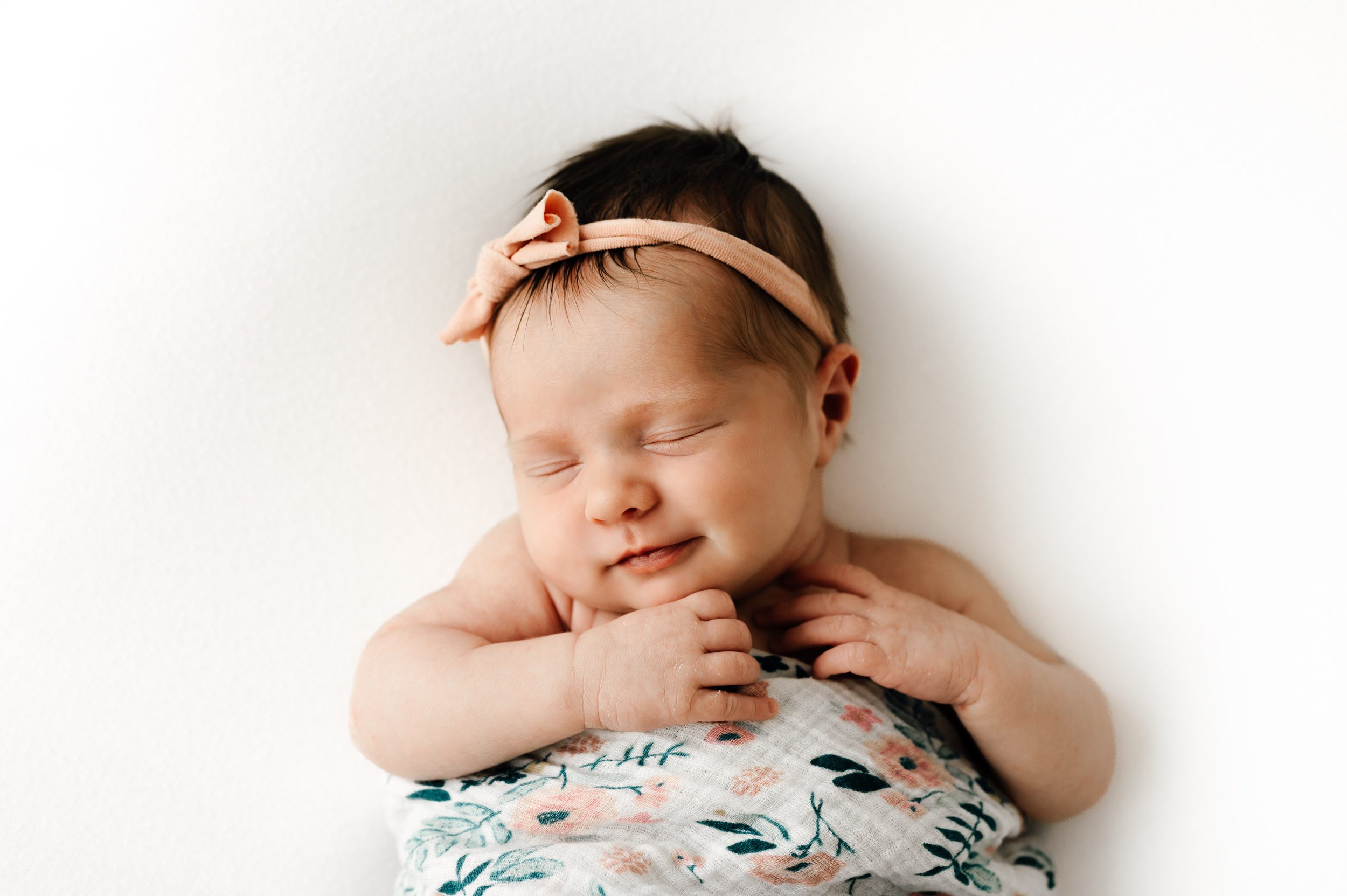 a baby girl wearing a pink headband wrapped in a floral swaddle laying on her back on a white backdrop with her hands tucked up under her chin and a hint of a smile on her face as she sleeps during a natural newborn photography session