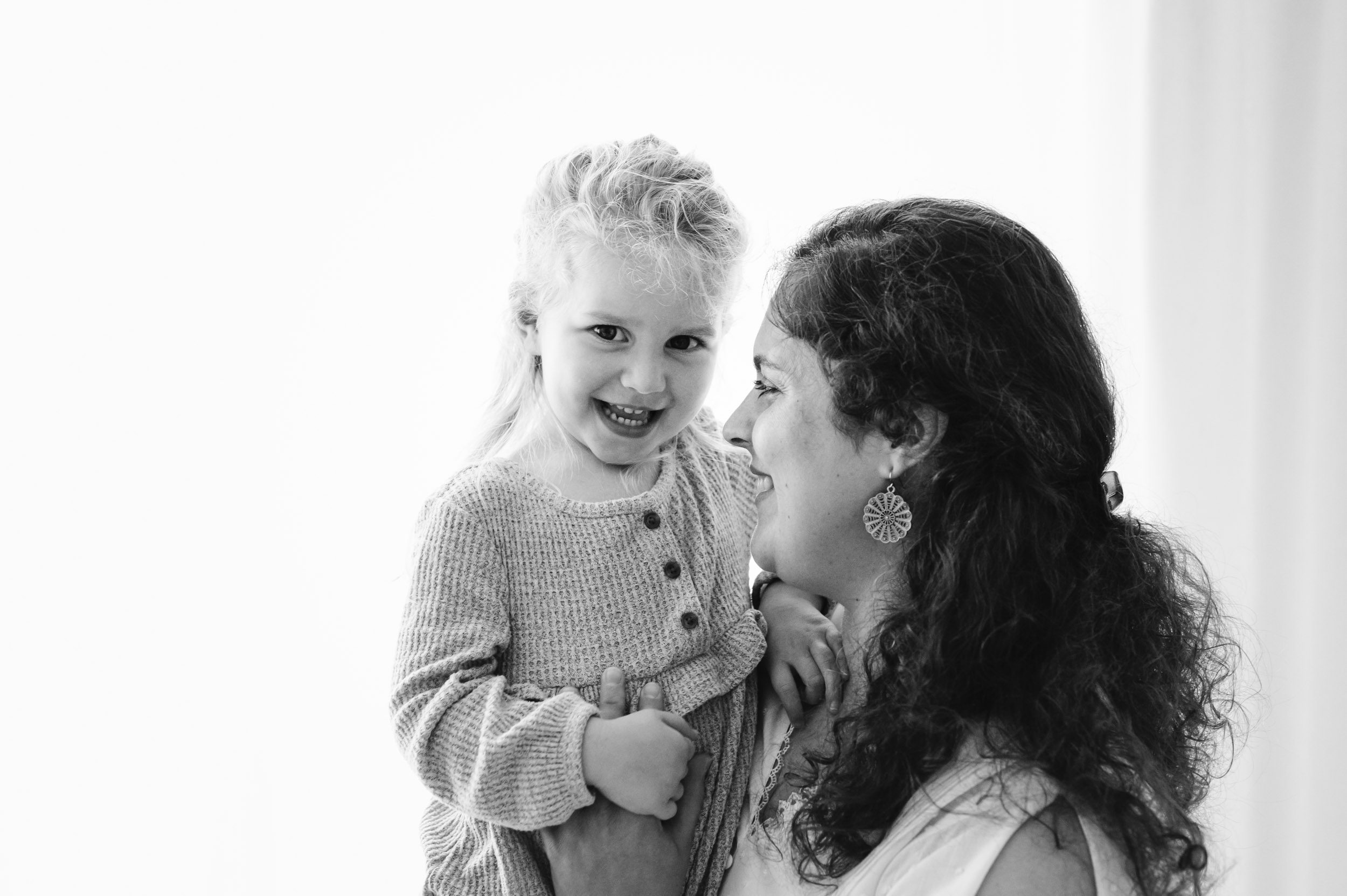 a black and white back lit picture of a mom holding her young daughter on her lap and smiling at her as she looks at the camera and laughs