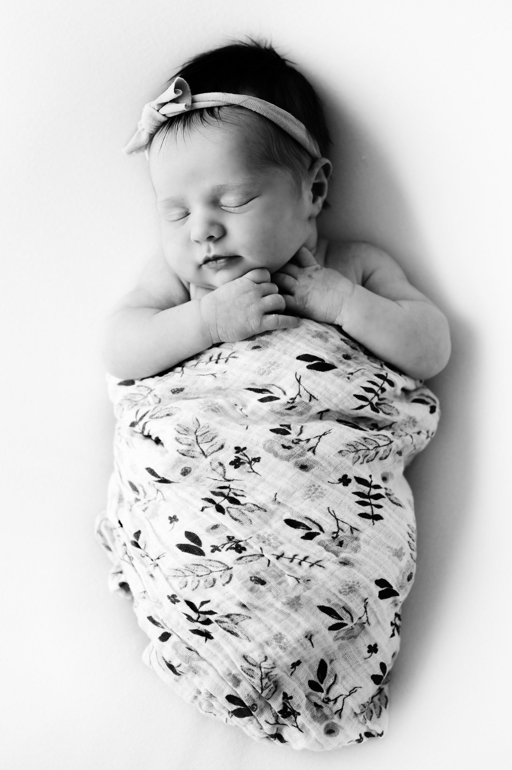 a black and white picture of a newborn baby girl laying on her back on a white backdrop wrapped in a floral swaddle blanket and tucking her hands under her chin as she sleeps during a natural studio newborn photography session