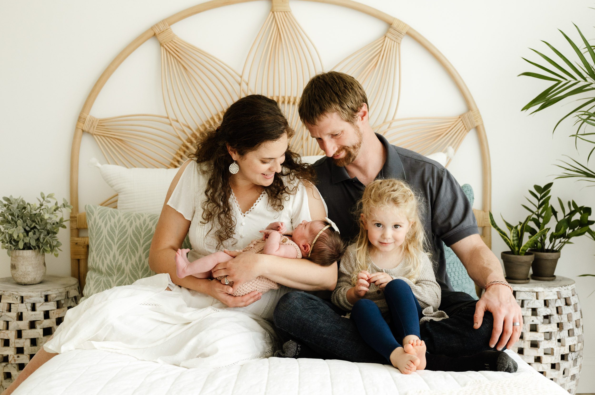 a new family of four sitting on a bed as mom and dad smile down at their baby girl cradled in mom's arms and big sister looks right at the camera with a shy smile on her face during a studio newborn photo session