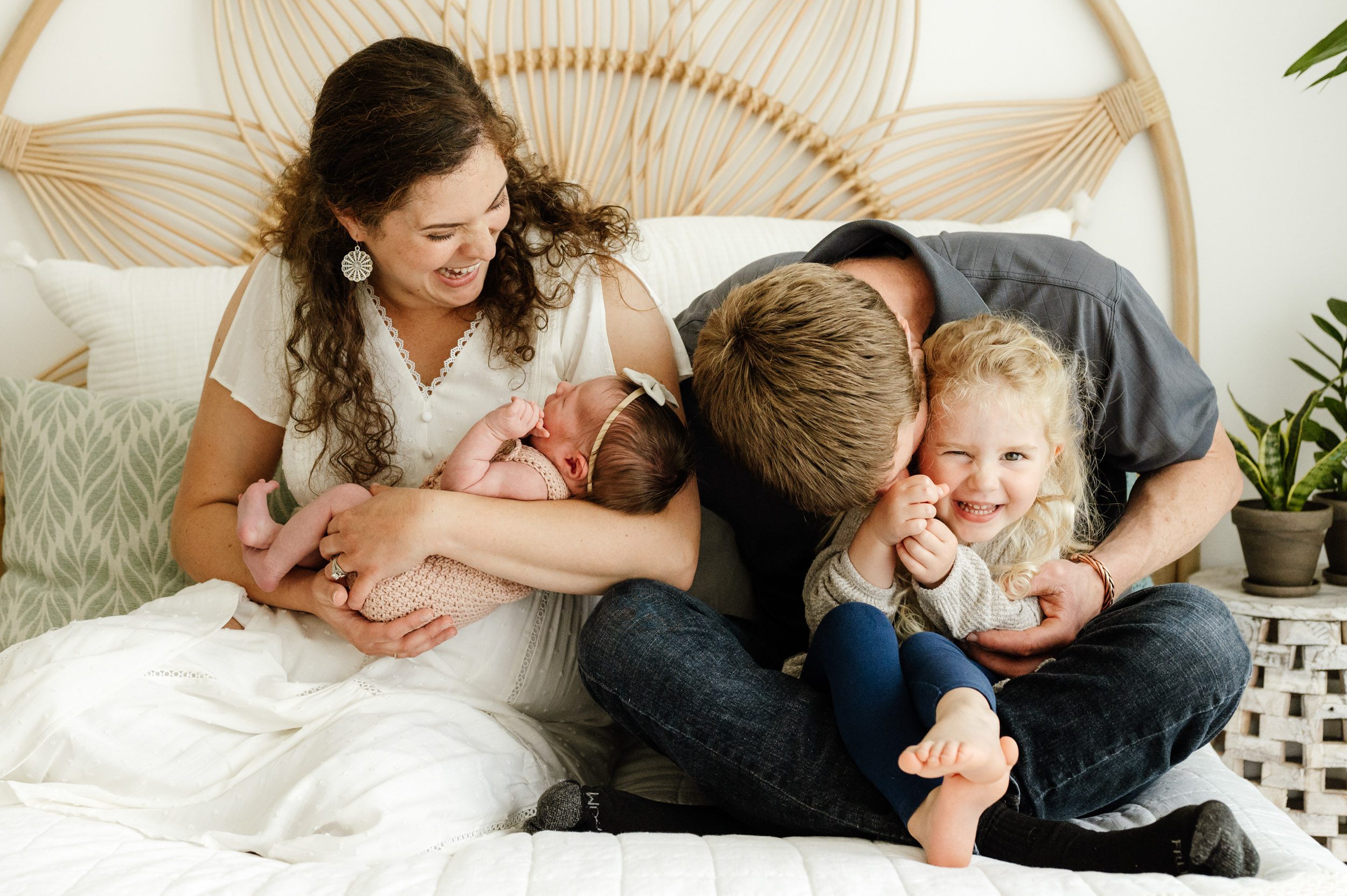 a new family of four sitting on a bed with the baby cradled in mom's arms and big sister sitting on dad's lap and laughing as dad leans down to blow raspberries on her cheek during a studio newborn photoshoot
