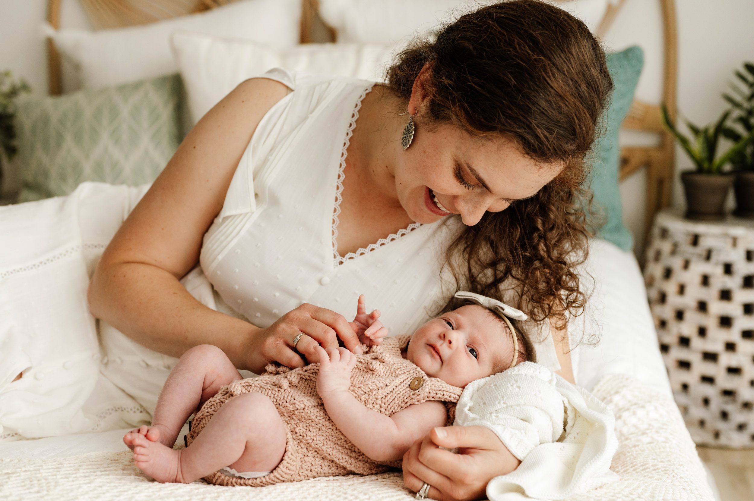 a new mom laying on a bed smiling down at her baby girl laying right in front of her as the baby looks toward the camera during a natural light studio newborn photo session