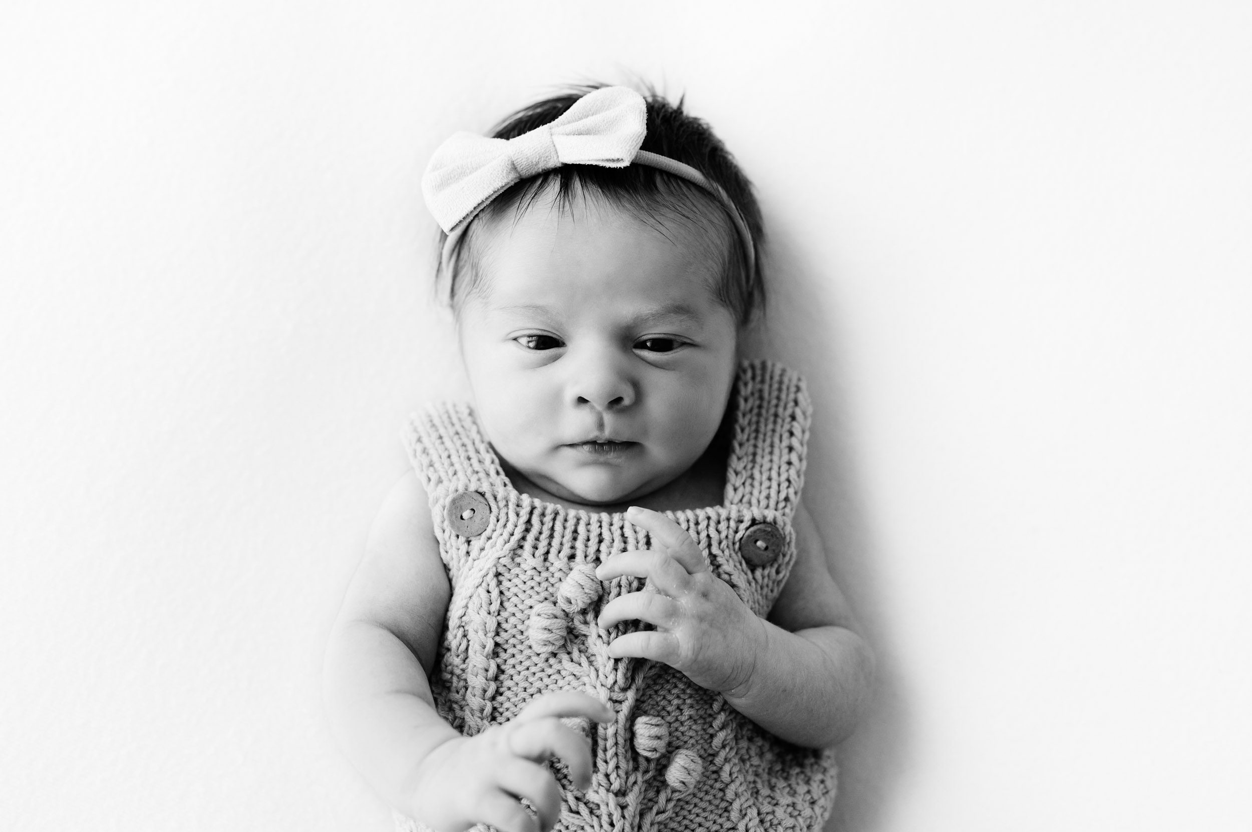 a black and white picture of a baby girl laying on her back on a white backdrop and looking right at the camera during a natural newborn photo session