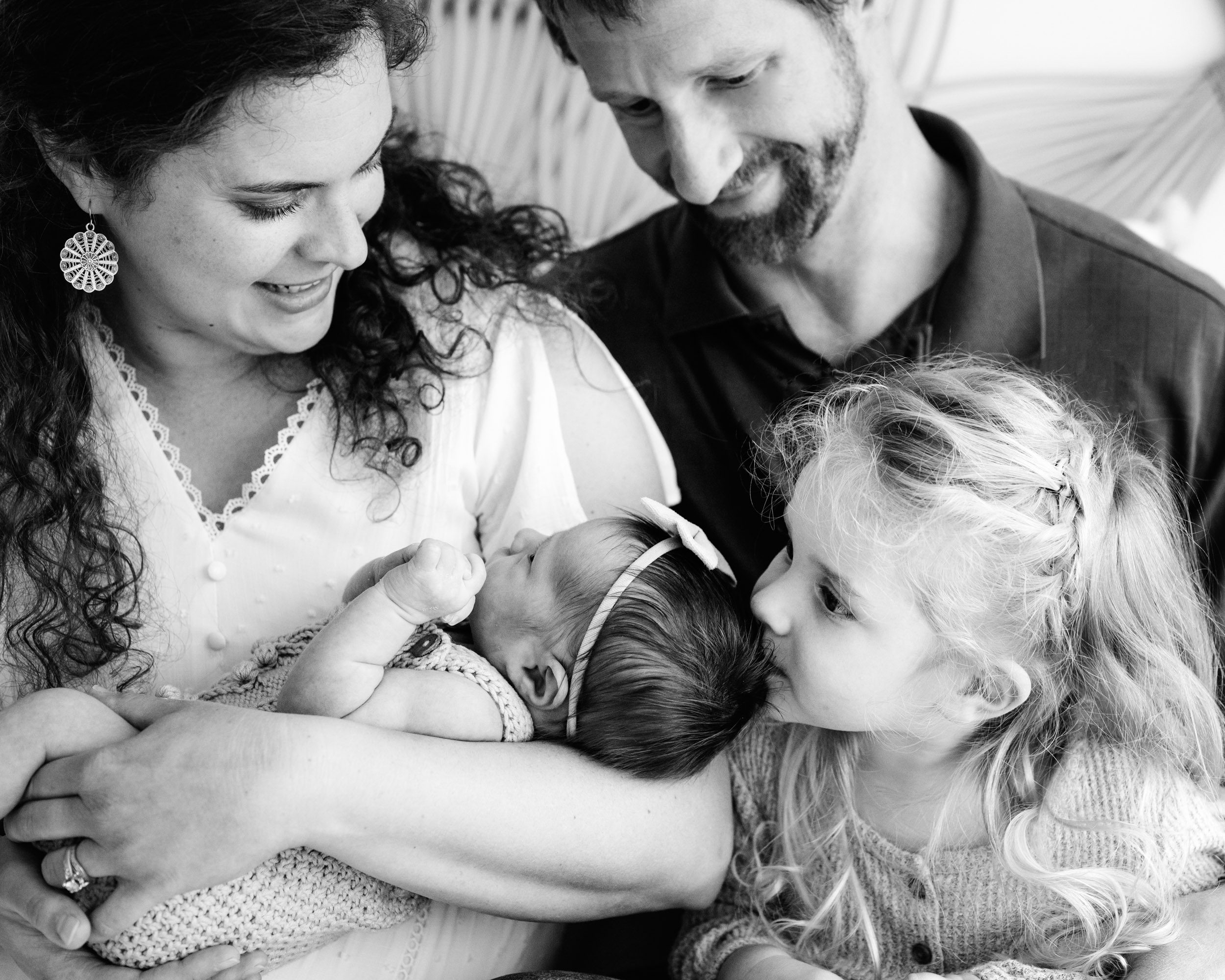 a black and white picture of a new family of four sitting on a bed while mom holds her newborn daughter cradled in her arms and big sister leans in to kiss her baby sister on the head during a natural newborn photography session in studio