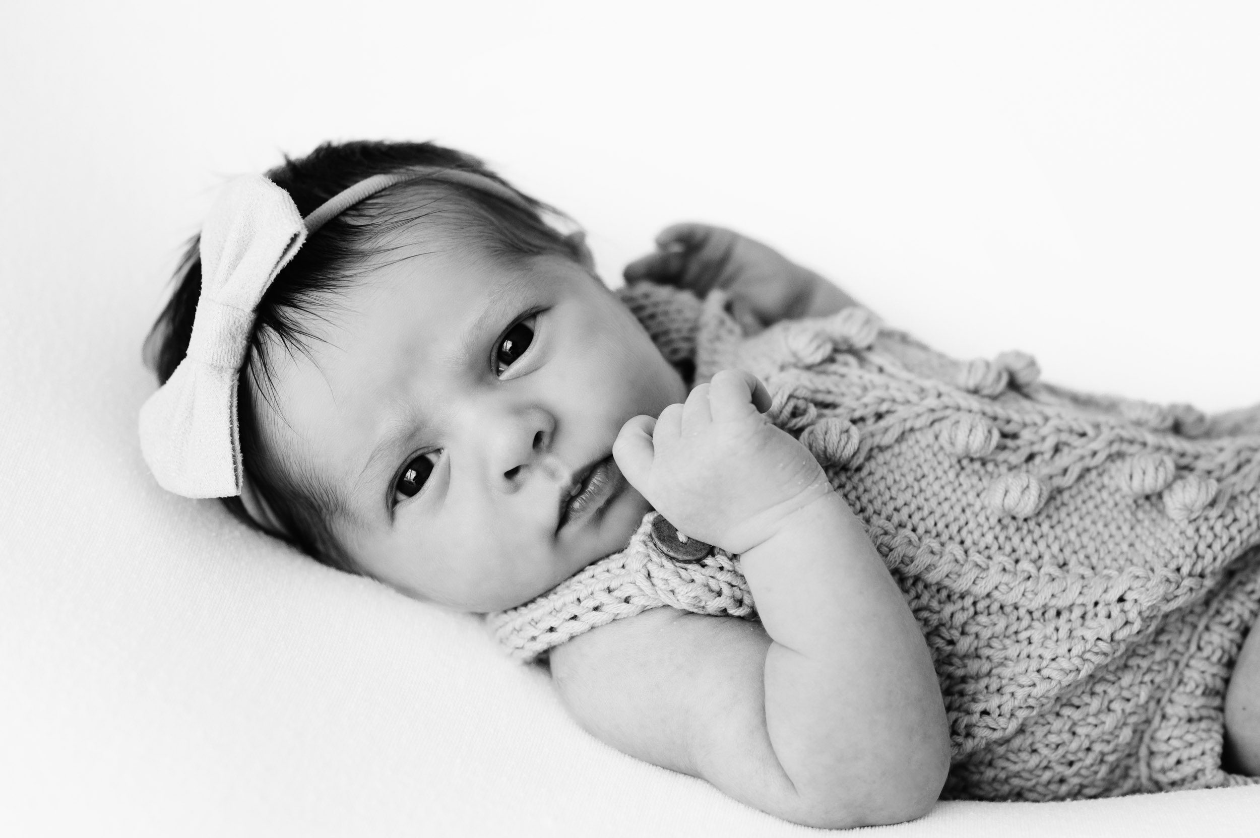 a baby girl in a knit onesie laying on a white background and touching her finger to her cheek as she looks directly at the camera during a studio newborn photoshoot