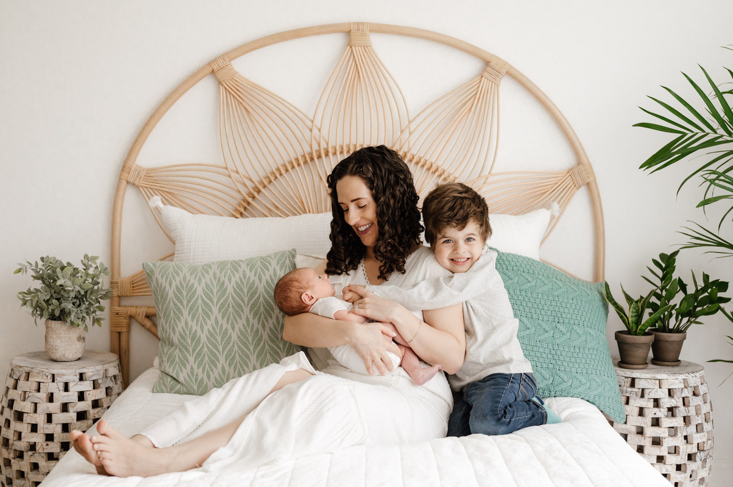 a mom sitting on a bed and smiling down at her newborn son cradled in her arms while her older son hugs her and smiles at the camera during a newborn photoshoot