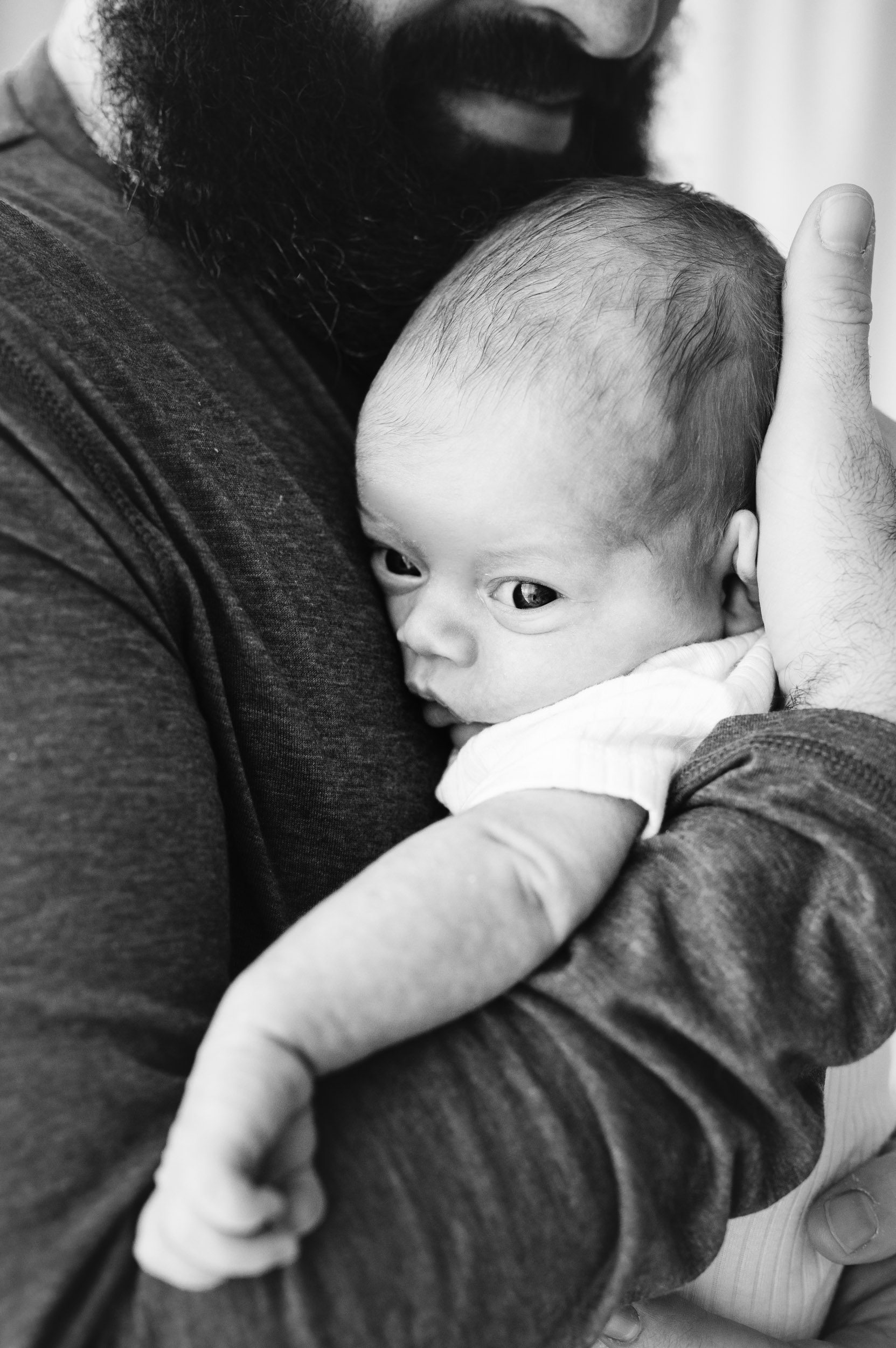 a black and white picture of a newborn baby boy snuggled up against his father's chest as he looks off to side toward the camera during a Pottstown studio newborn session