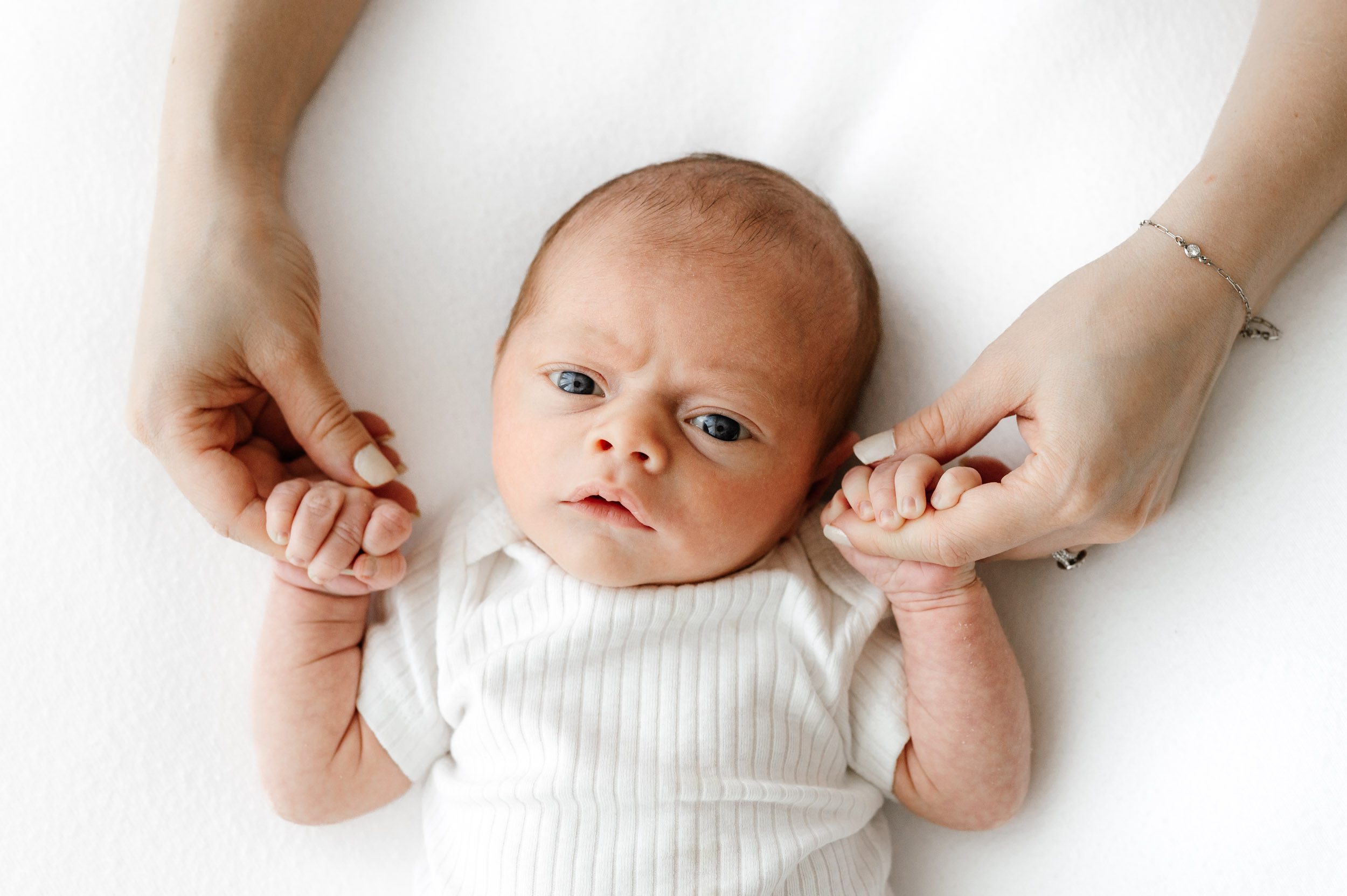 a baby boy laying on a white backdrop and gazing up at the camera as he grabs onto his mom's fingers during a philadelphia newborn photo session