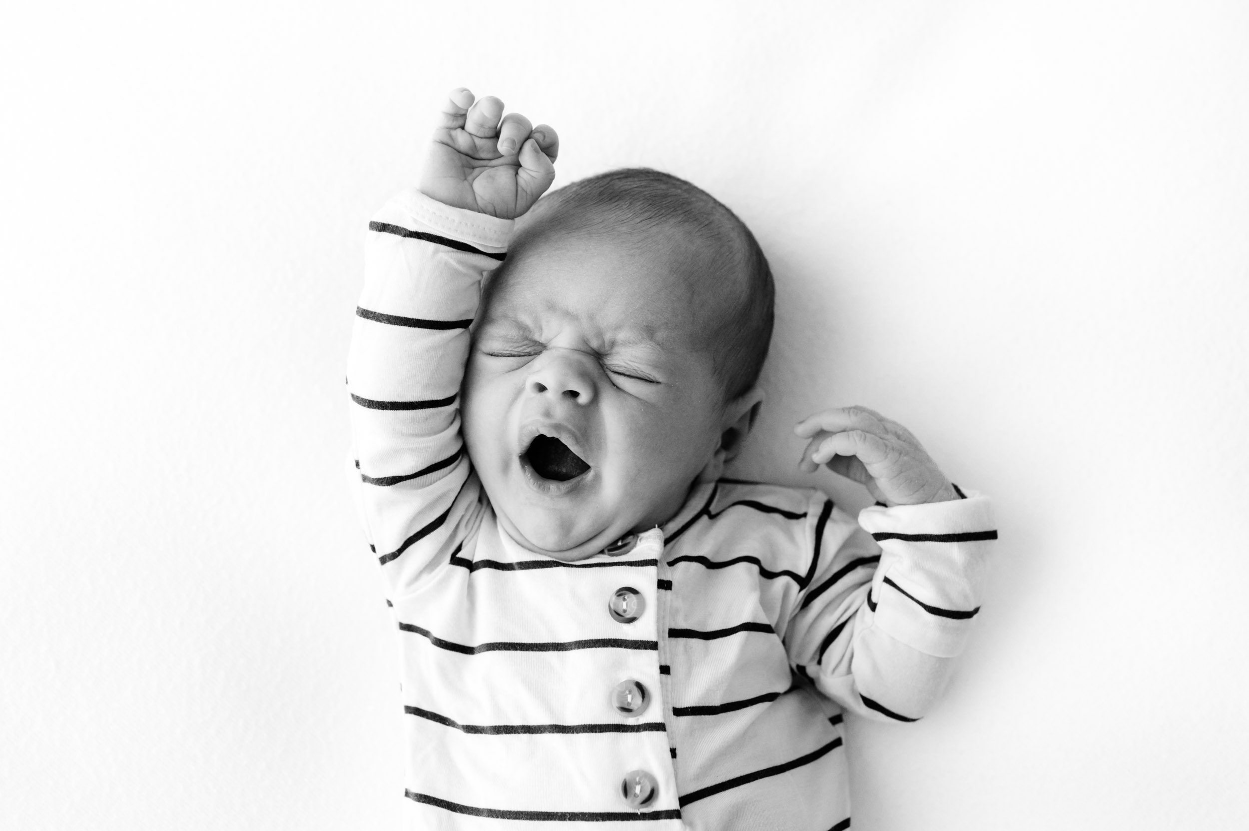 a black and white picture of a baby boy in a white and black striped romper laying on a white backdrop as he stretches and yawns during a Philadelphia newborn photo session