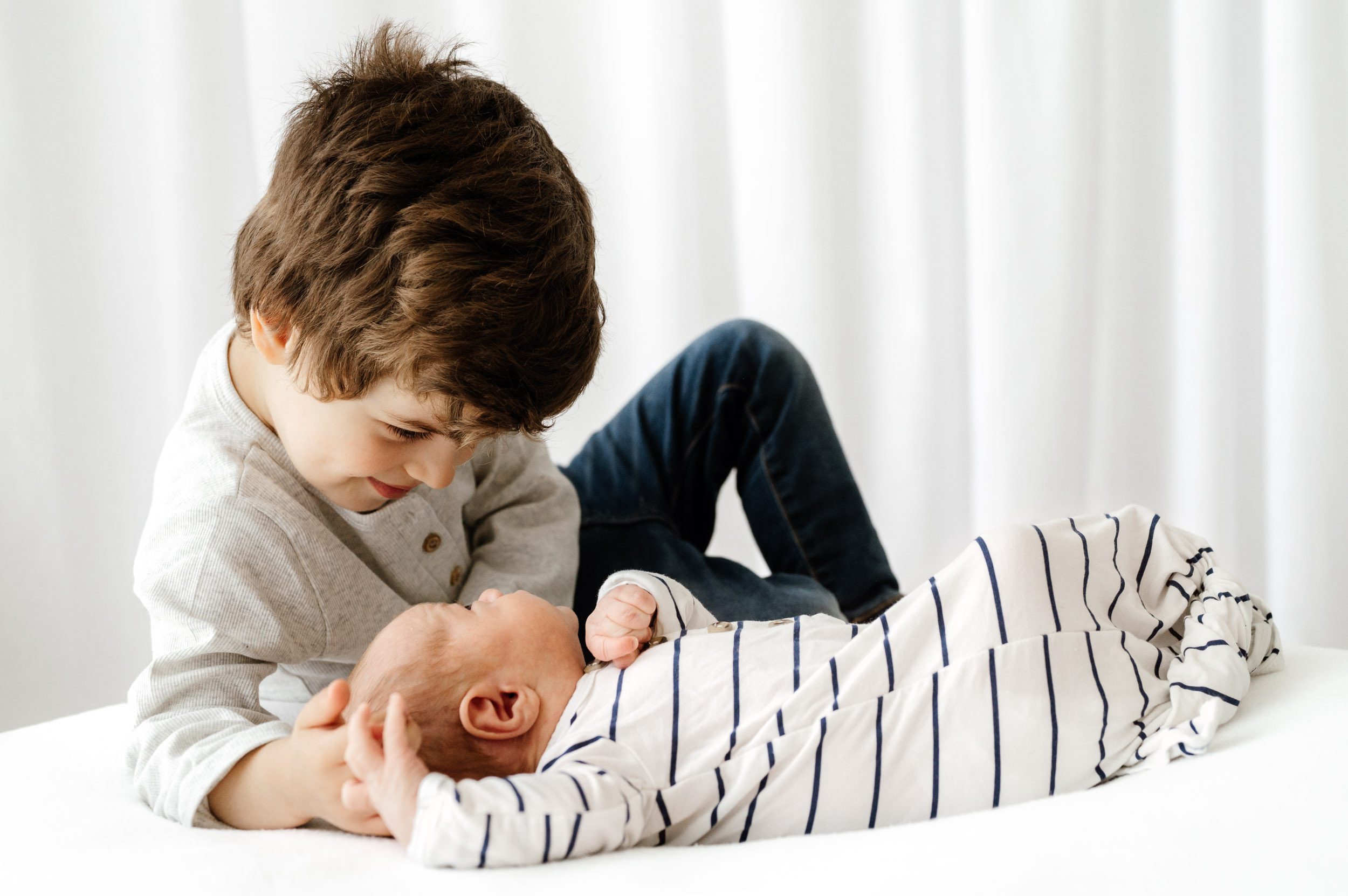 a baby boy laying on a white bean bag  and looking up at his older brother as he touches the baby's head and smiles down at him during a Pottstown studio newborn photo session