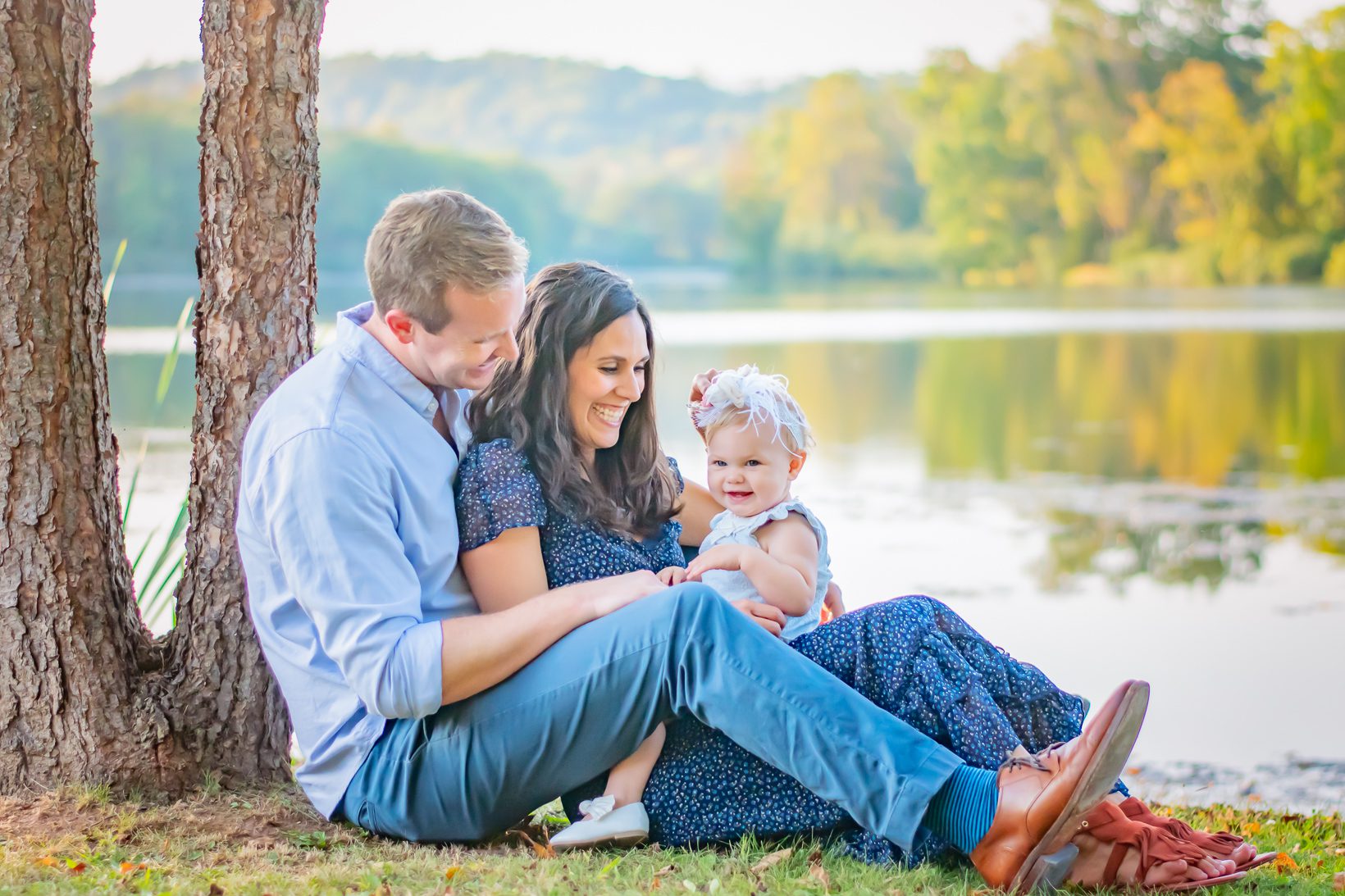 a family of three sitting in front of a lake with their young daughter on their lap and the colorful leaves reflecting off the water in the background during a fall mini session