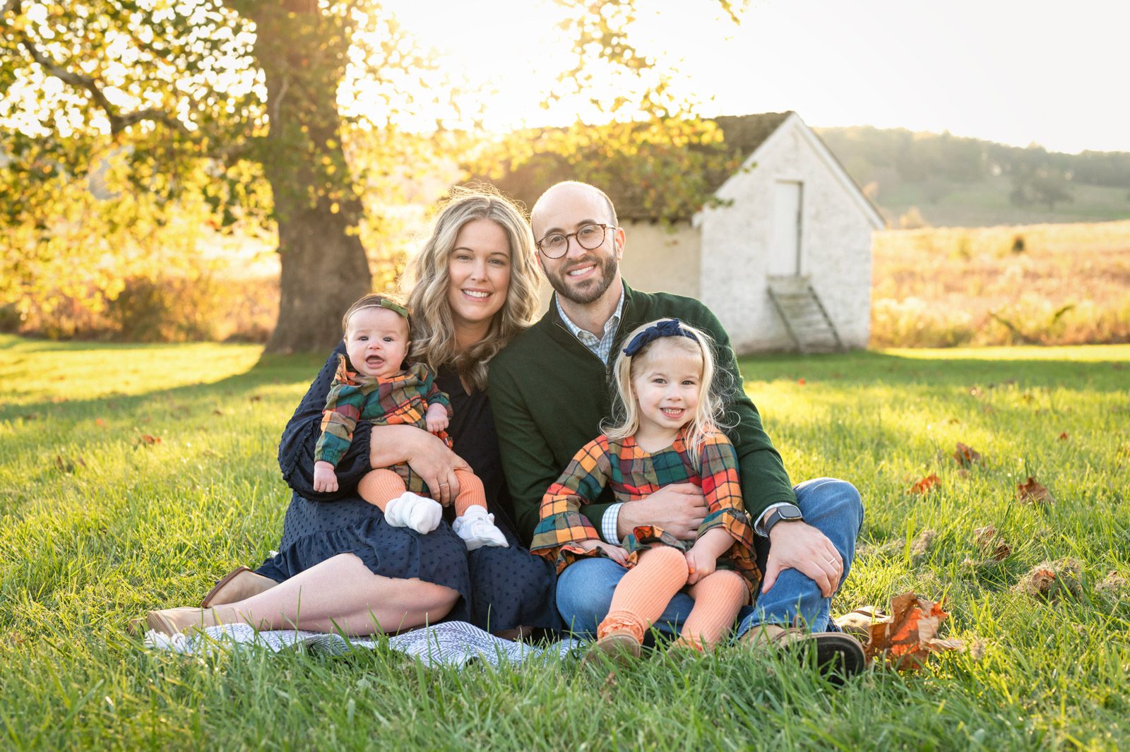 a mom and dad sitting in the grass with their two young daughters on their lap as everyone smiles at the camera while the sun shines through the colorful leaves in the background during a fall mini session