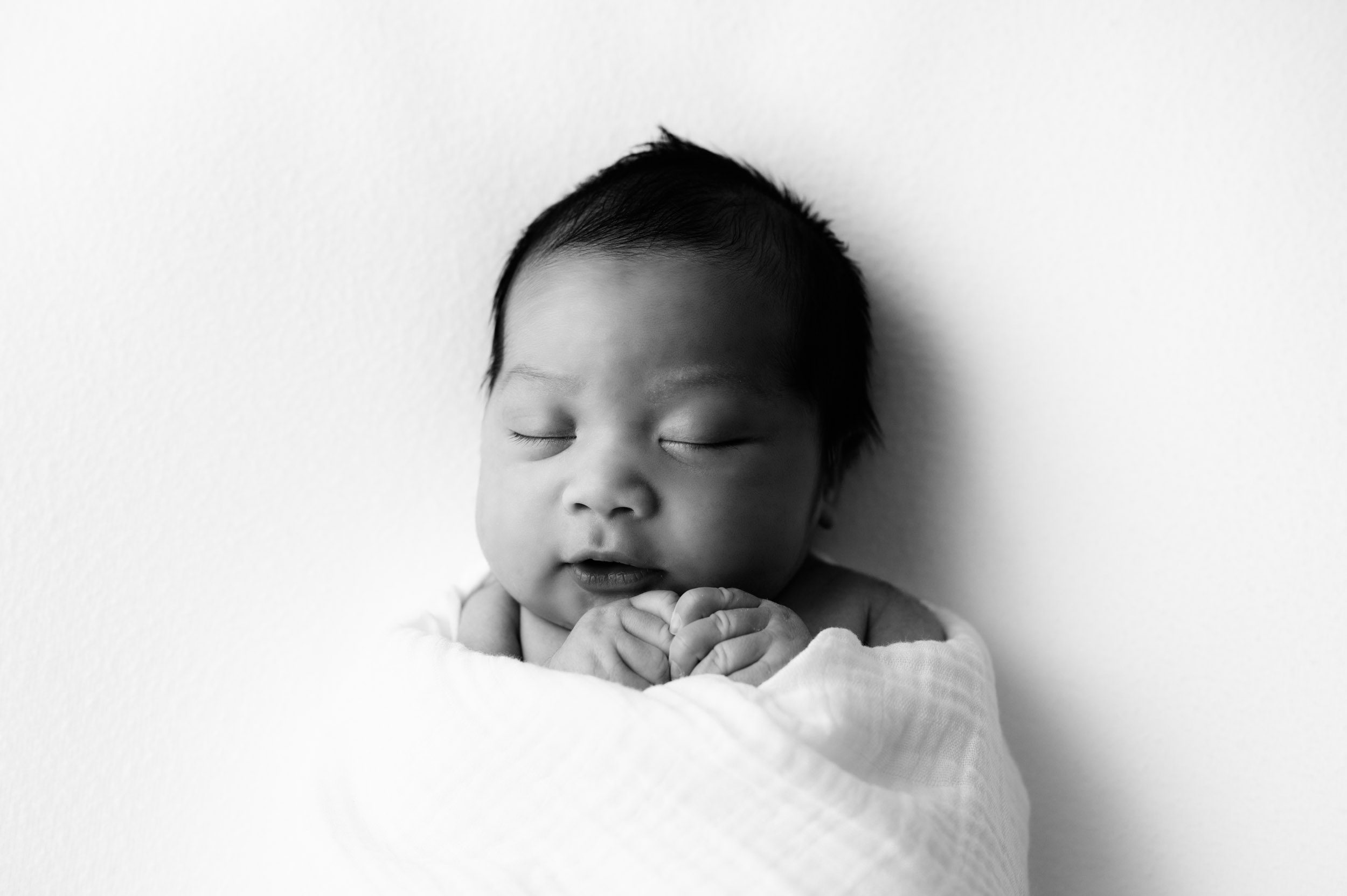 a black and white picture of a baby boy wrapped in a white swaddle laying on a white backdrop sleeping with his hands tucked up under his chin during a newborn photoshoot