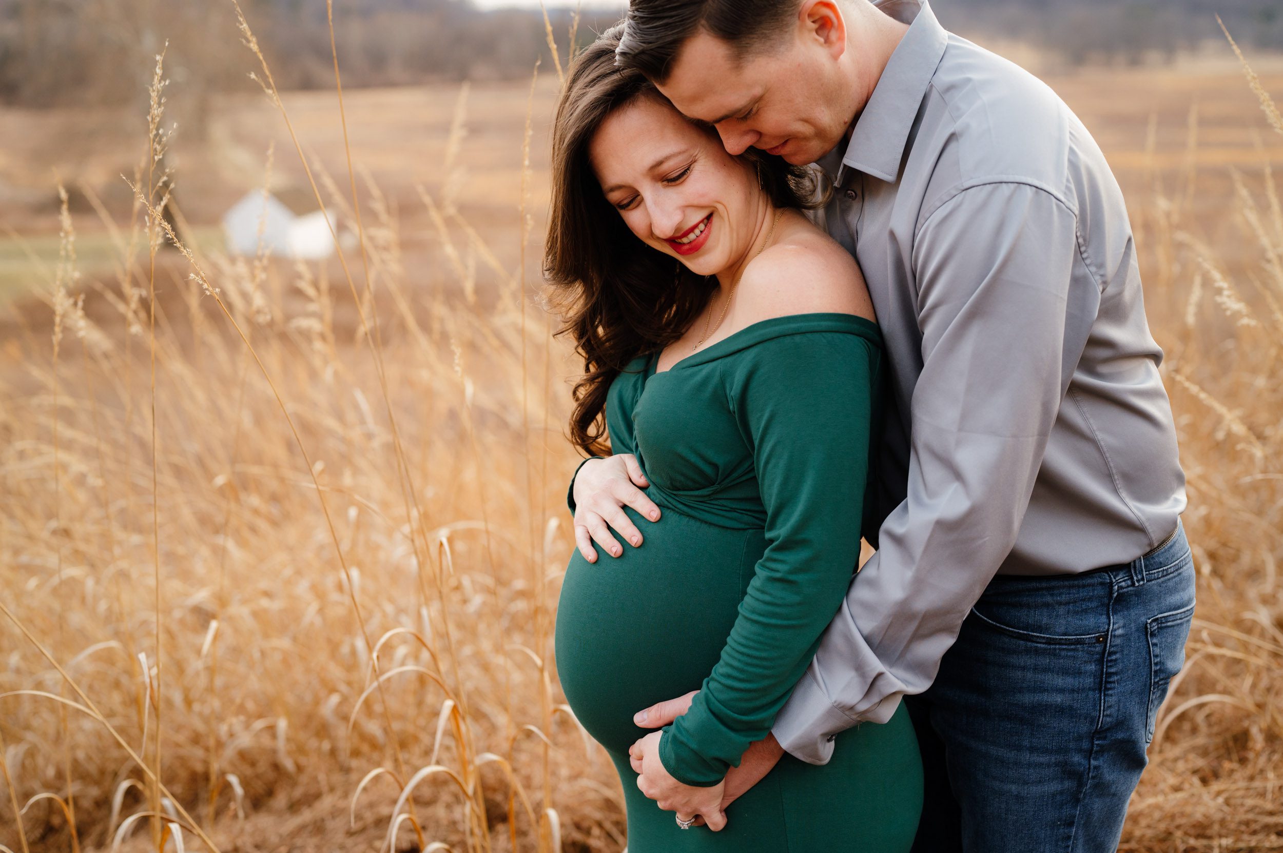 an expecting mom standing in a field of golden grasses as her husband hugs her from behind and they both smile down at her belly during a maternity photo session