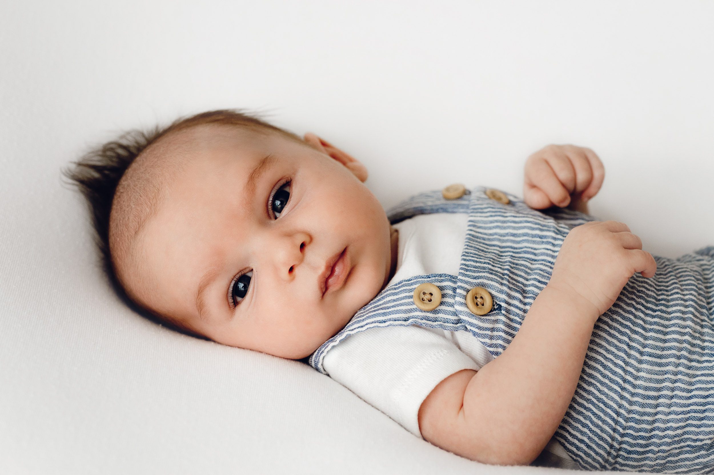 an 8 week old baby boy with mohawk hair wearing a blue and white striped romper laying on a white backdrop and looking directly at the camera during a newborn photo session