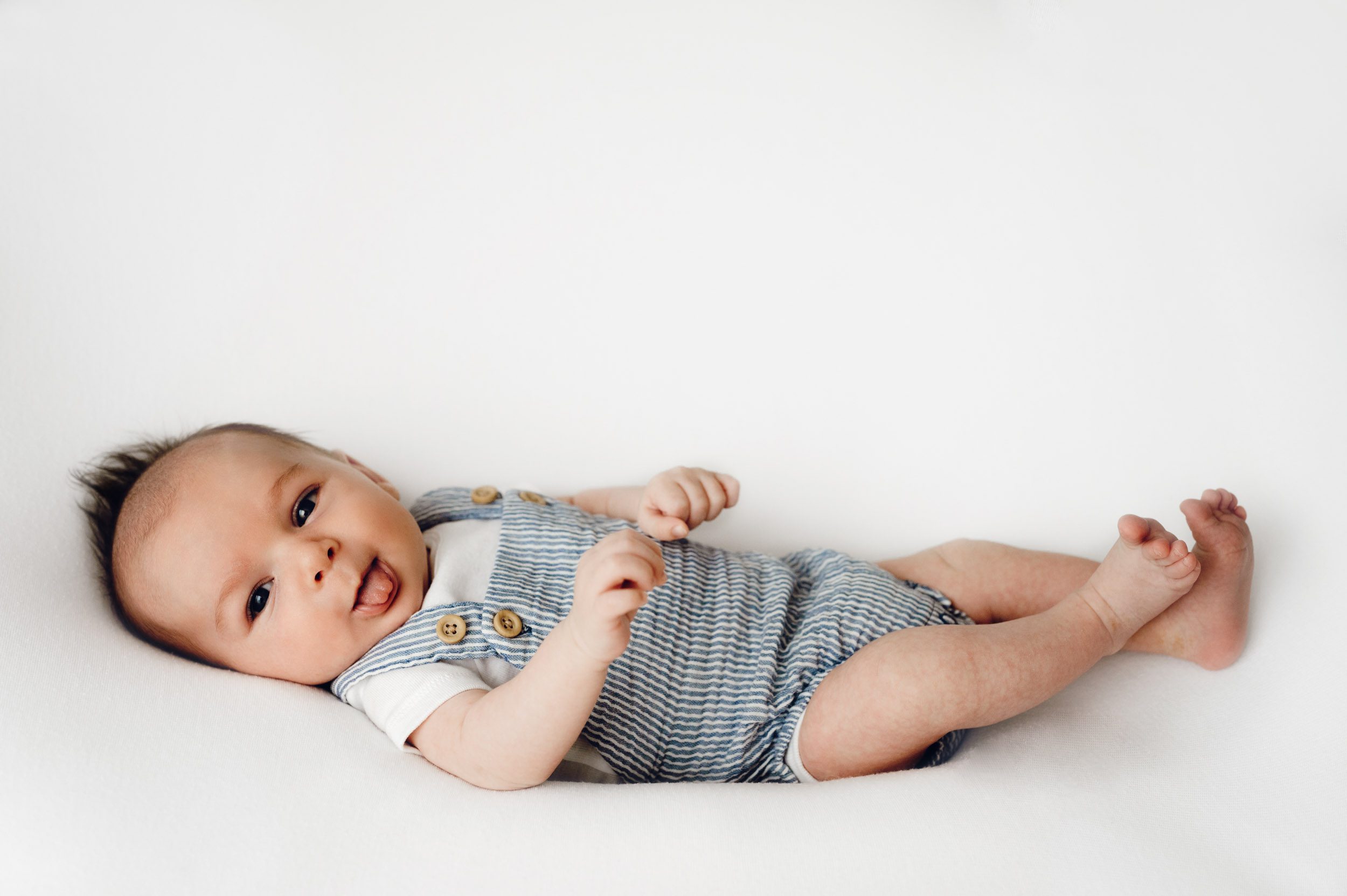 an 8 week old baby boy wearing a blue and white striped romper laying on a white backdrop and sticking his tongue out during a newborn photos session