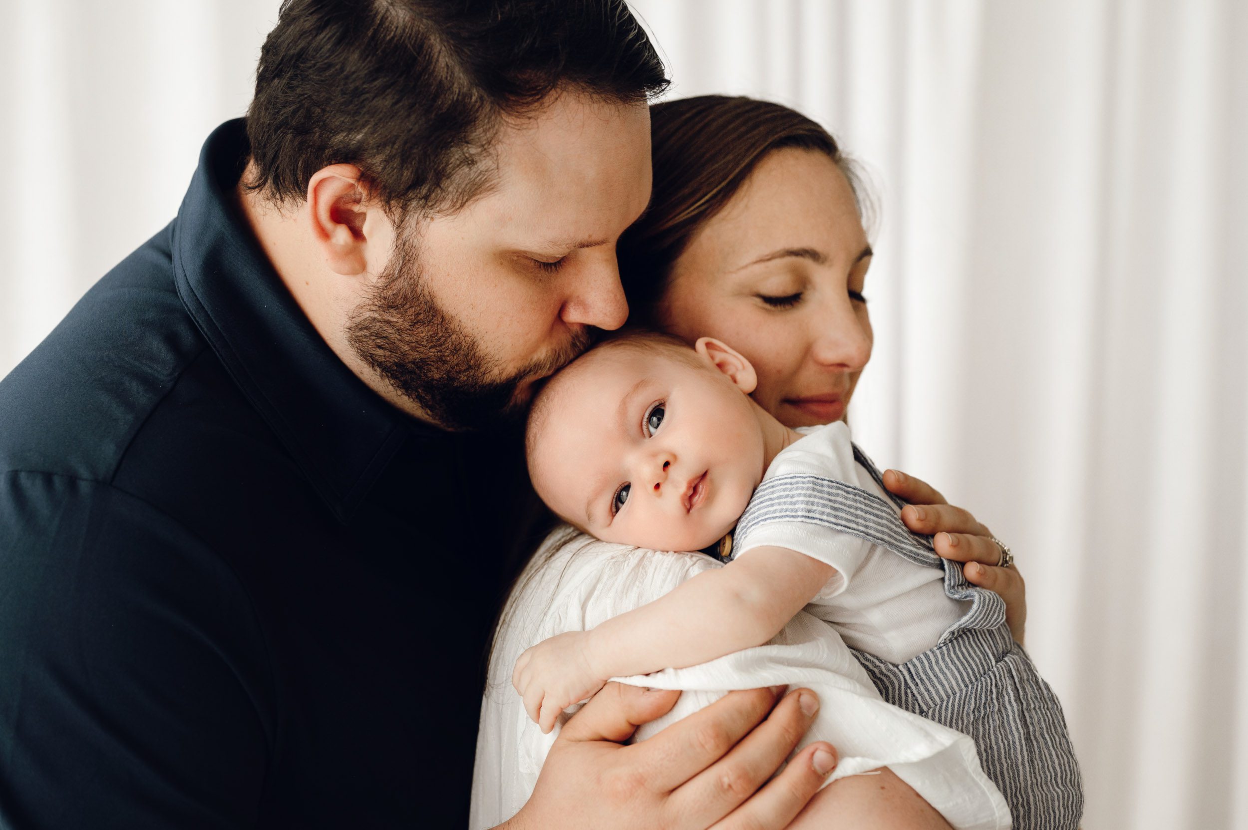 a new mom holding her baby boy against her chest as dad hugs her from behind and kisses his sown on the head while the baby looks directly at the camera during a newborn photos session