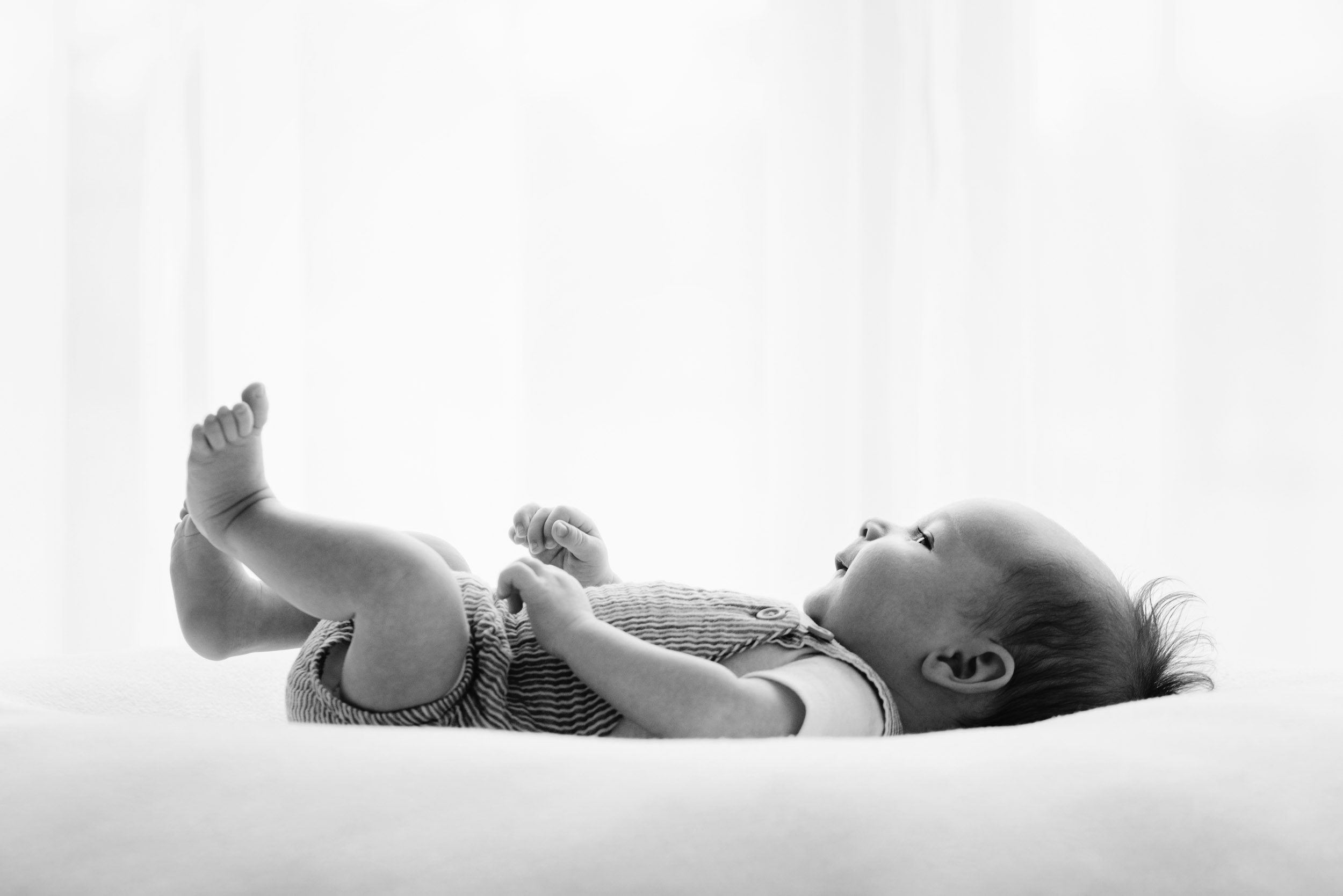 a black and white backlit profile picture of an 8 week old baby boy laying on a white bean bag and lifting his legs in the air as he smiles up at his mom during a newborn photos session