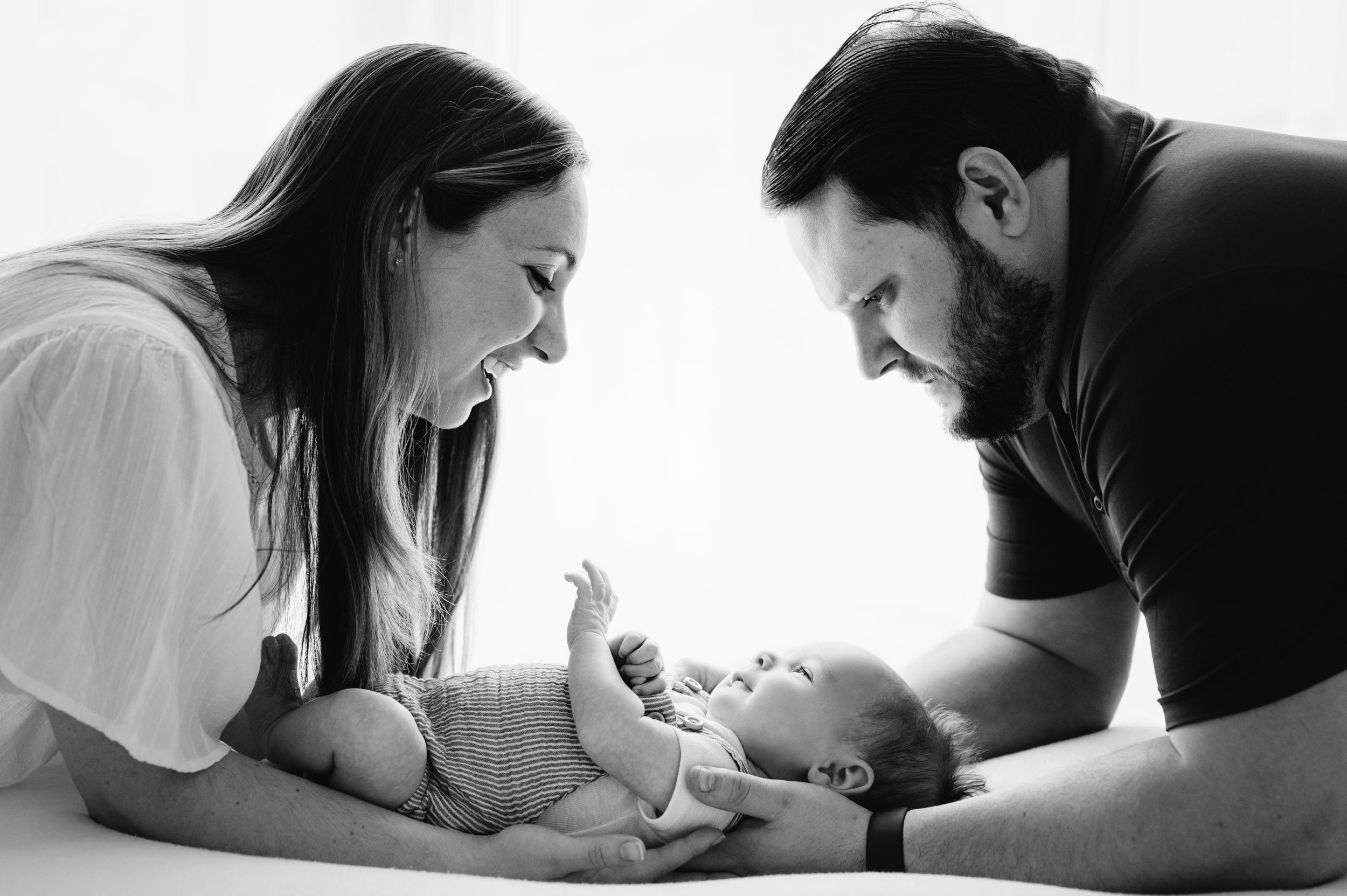 a black and white backlit picture of a baby boy laying on a white bean bag with his parents kneeling on either side of him with their arms around him as they smile down at him during a newborn photos session