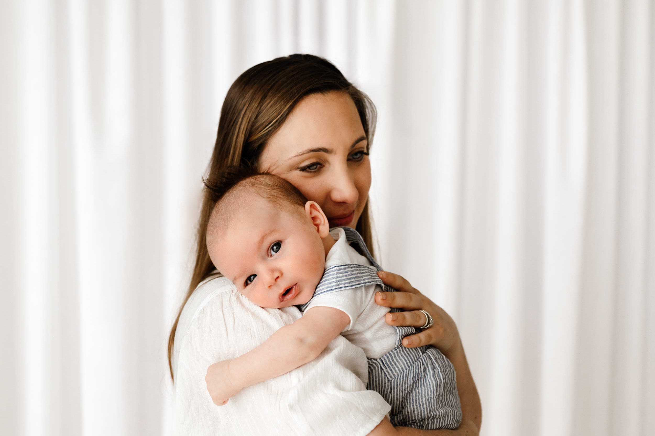 a new mom standing with her baby boy cradled against her as he rests his head on her shoulder and gazes directly at the camera during a newborn photoshoot