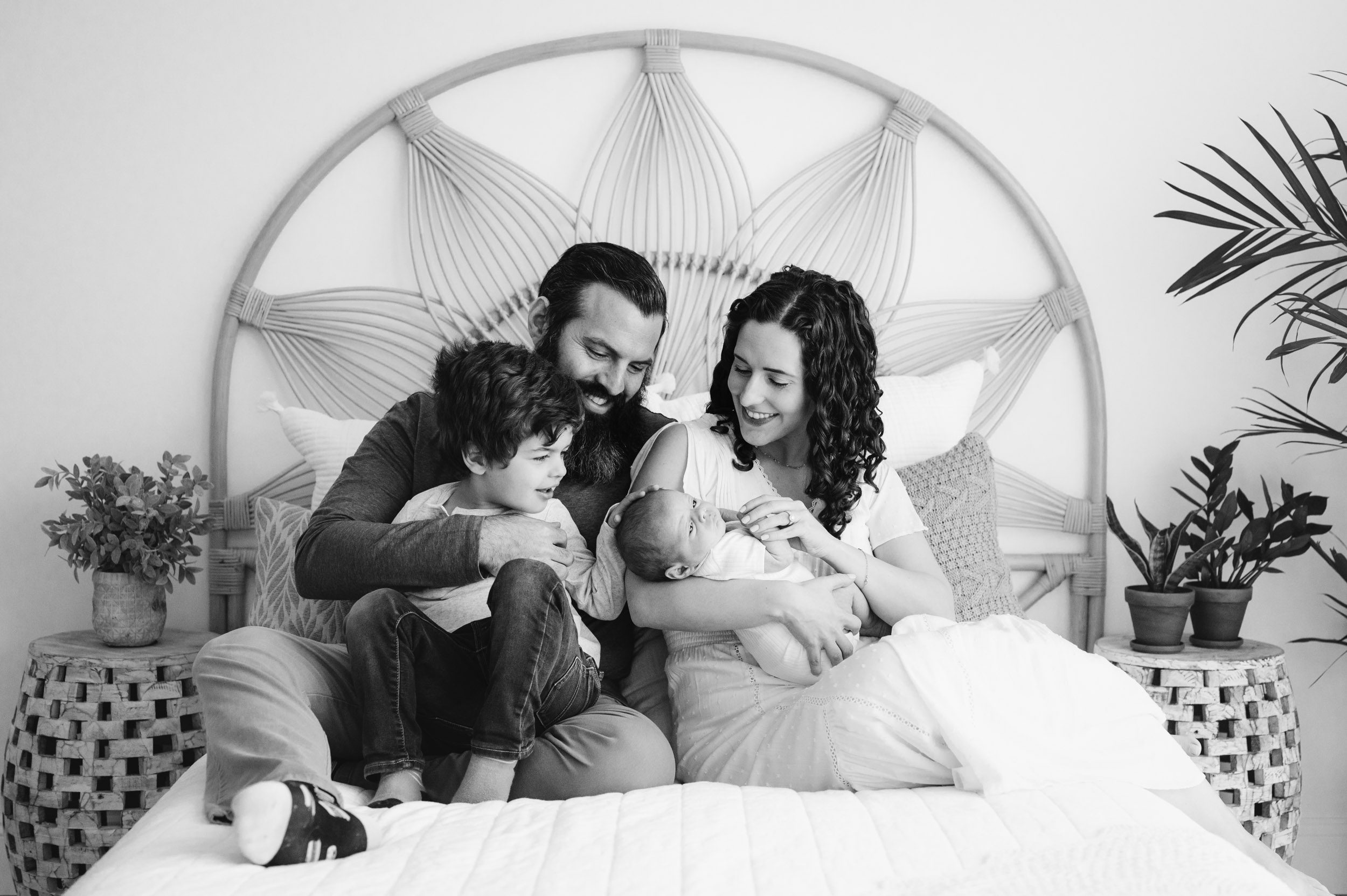 a black and white picture of a new family of four sitting on the bed while the parents smile down at the baby and big brother gently touches his baby brother on the head during a newborn photos session