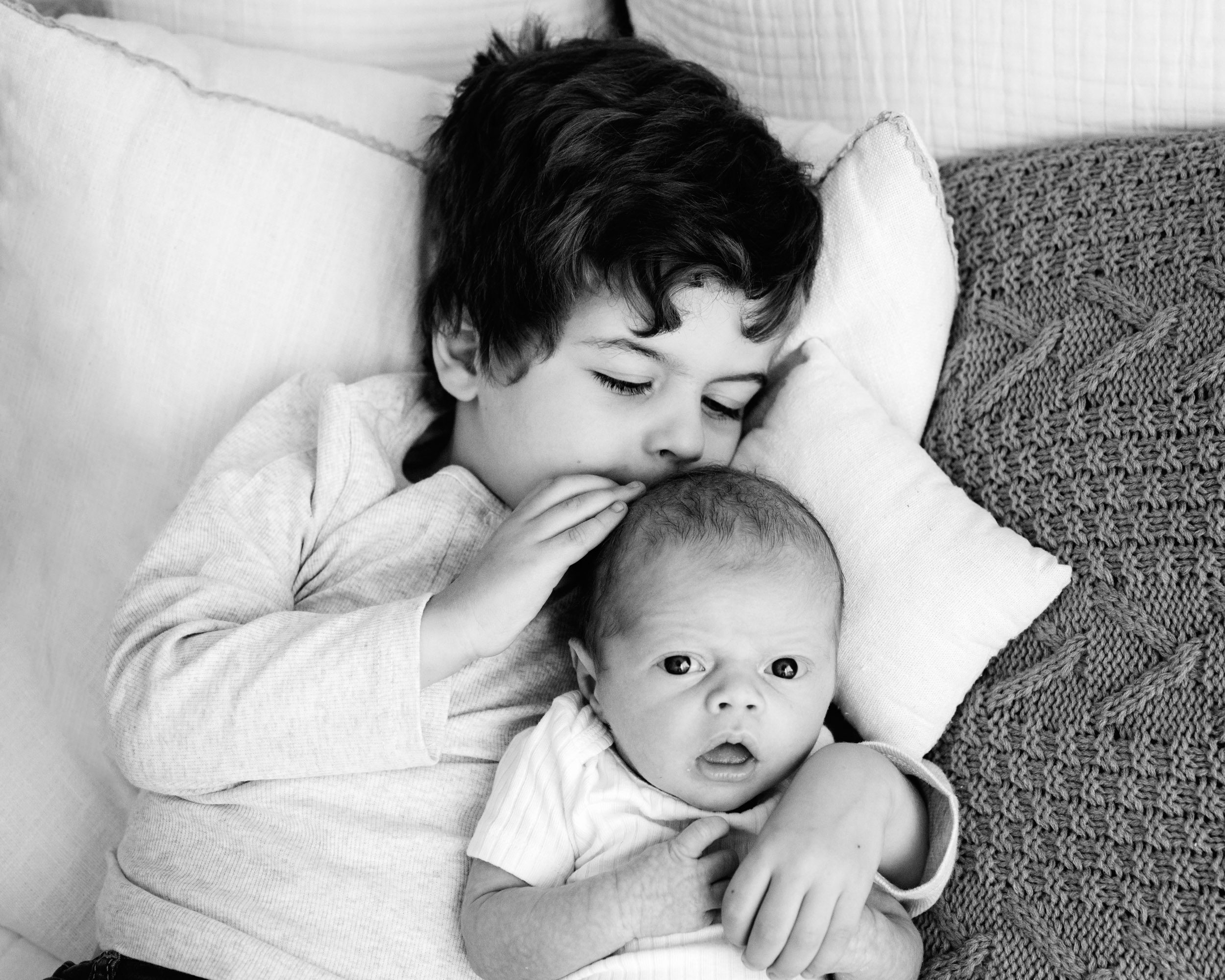 a black and white picture of a big brother laying on a bed and kissing his baby brother on the head while the baby looks directly at the camera during a newborn photoshoot