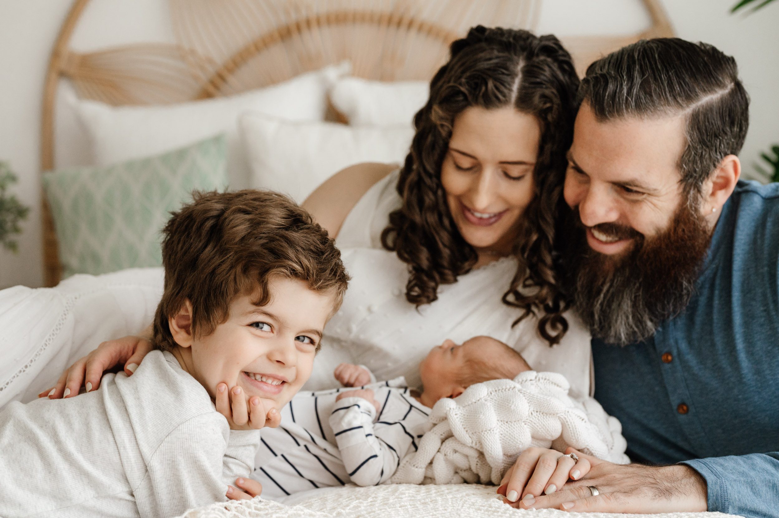 a new family of four laying on a bed with their baby boy as big brother looks directly at the camera with a huge smile on his face during a philadelphia newborn photography session