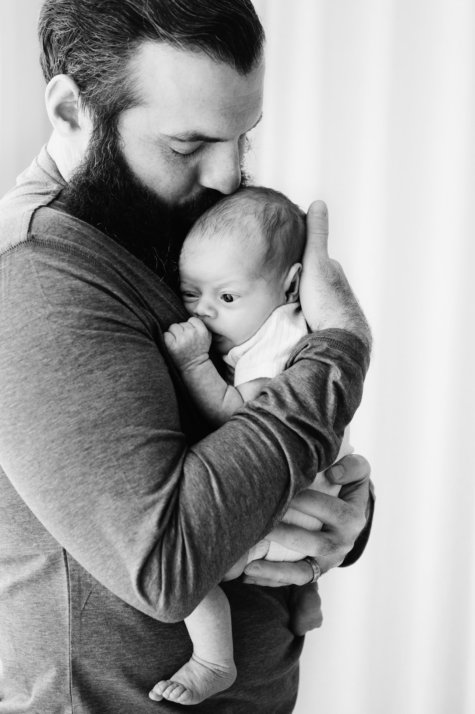 a black and white picture of a new father holding his baby boy against his chest and kissing him on top his head during a Philadelphia newborn photography session