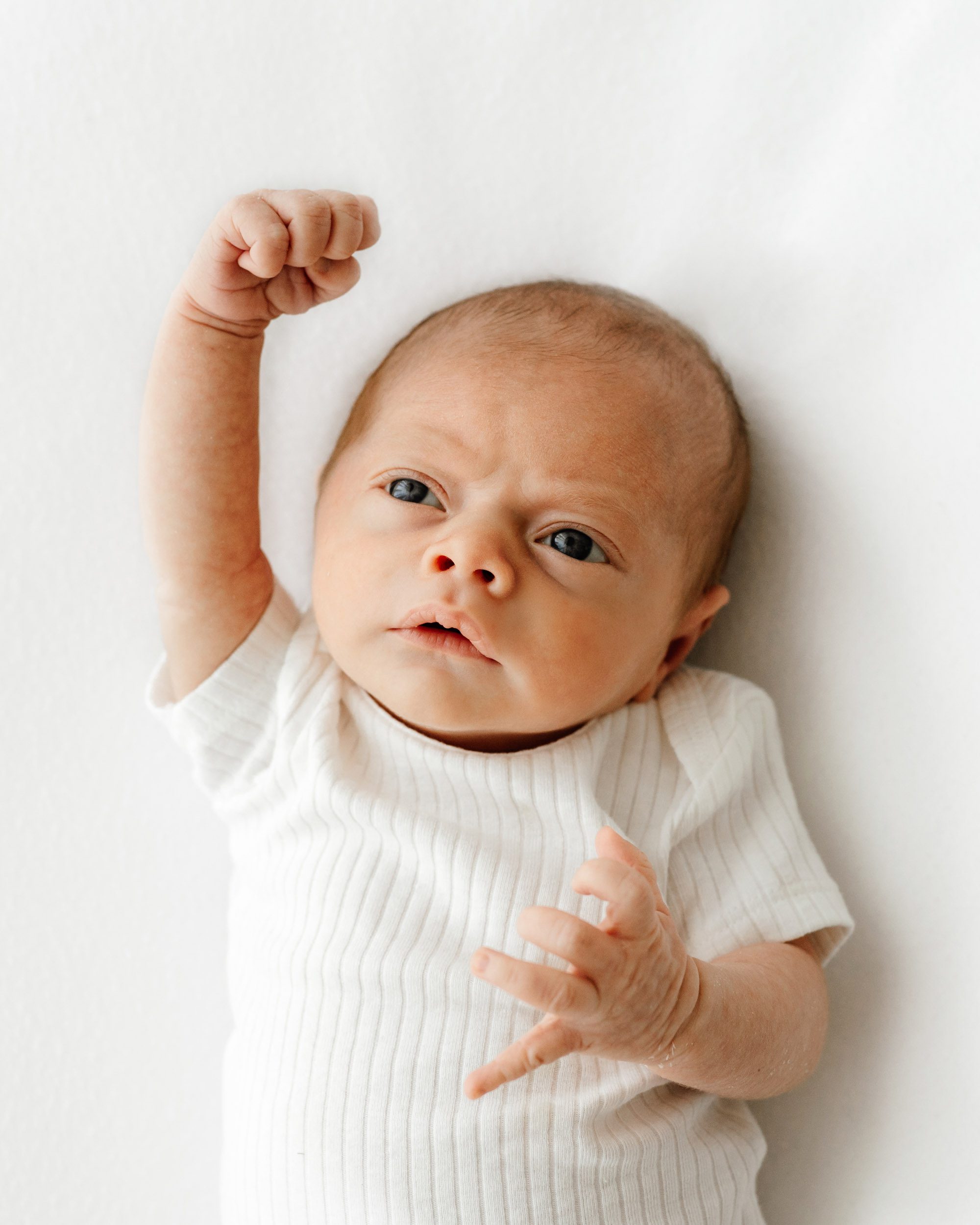 a baby boy in a white ribbed onesie laying on a white backdrop lifting his fist into the air above his head as he looks up toward the window light during a natural light studio newborn photoshoot