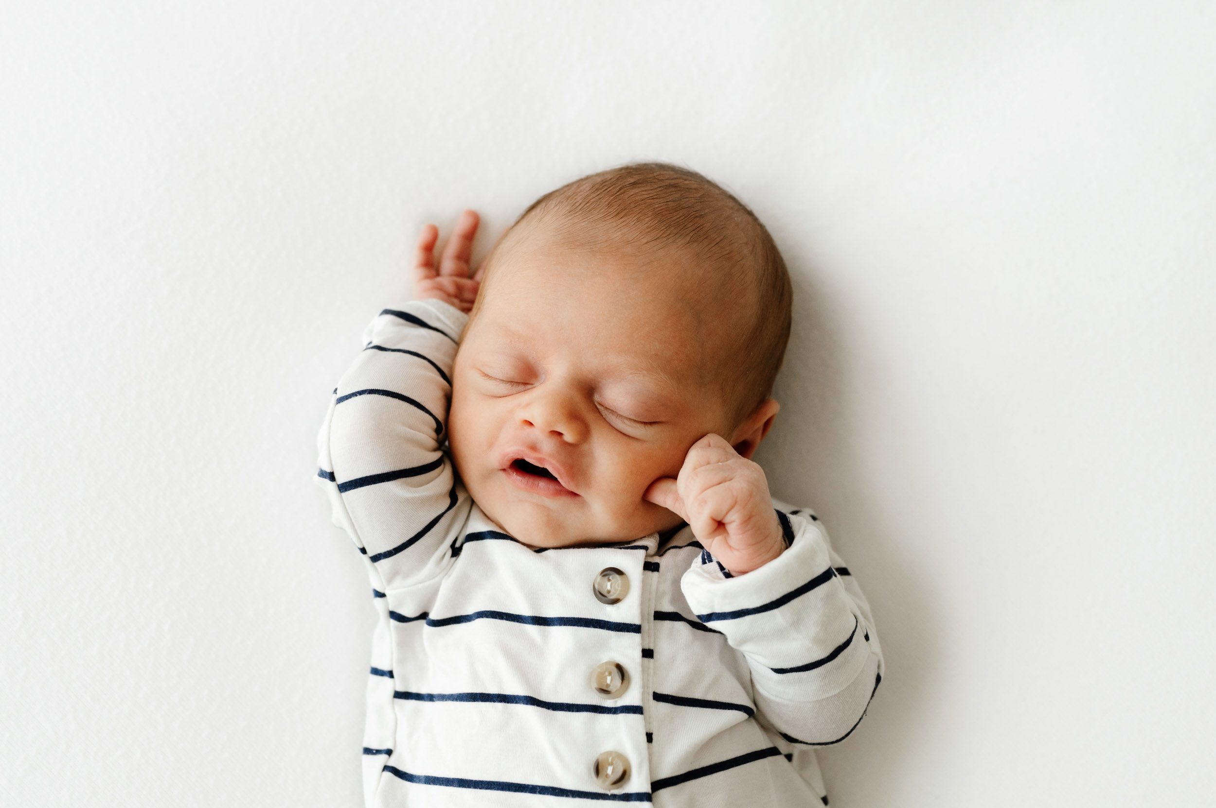 a baby boy wearing a blue and white striped romper laying on a white backdrop and holding his hands up next to his face during a Philadelphia newborn photoshoot