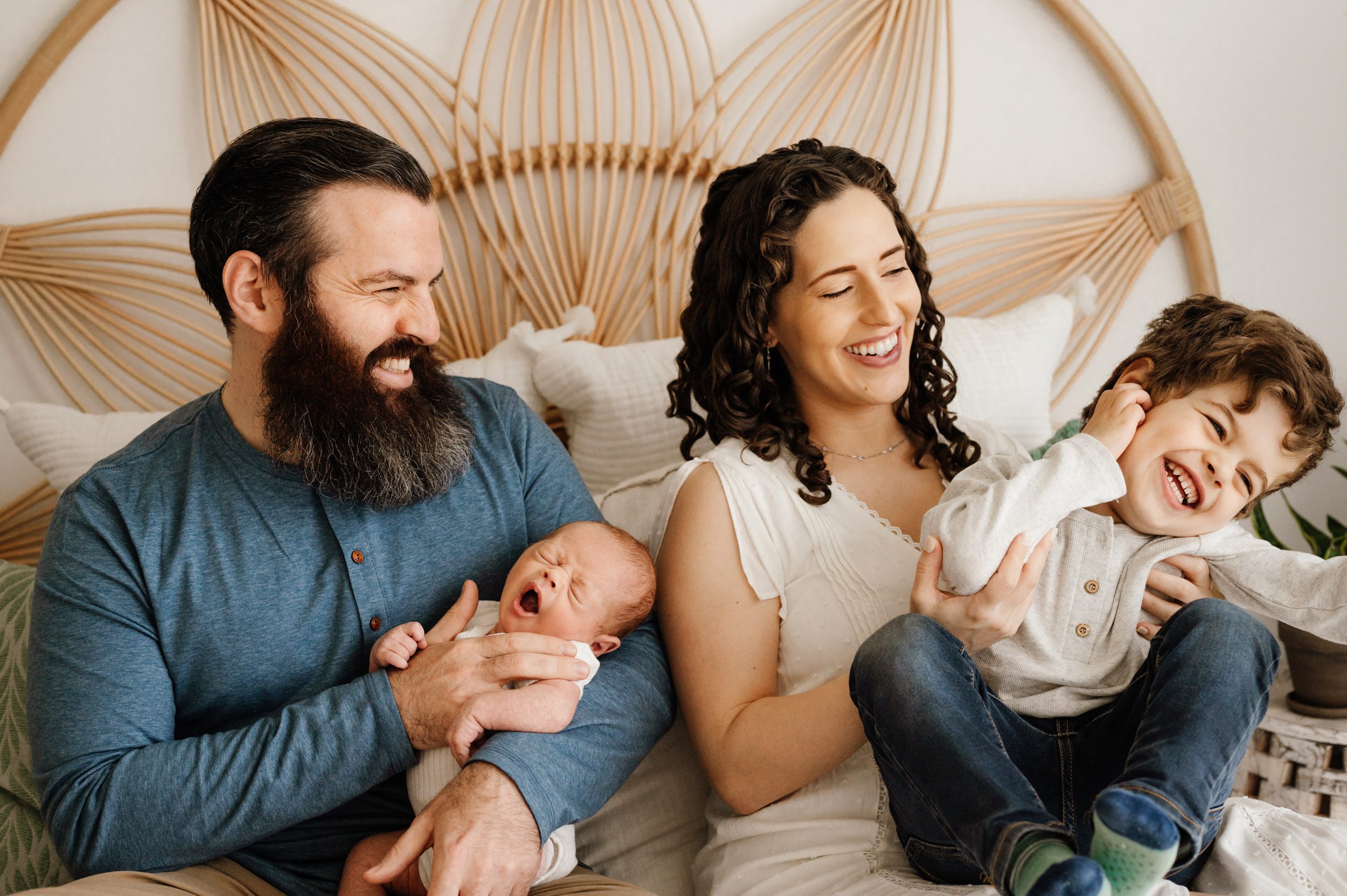 a new family of four sitting on a bed while mom and dad tickle and laugh at big brother and the baby yawns in dad's arms during a newborn photography session