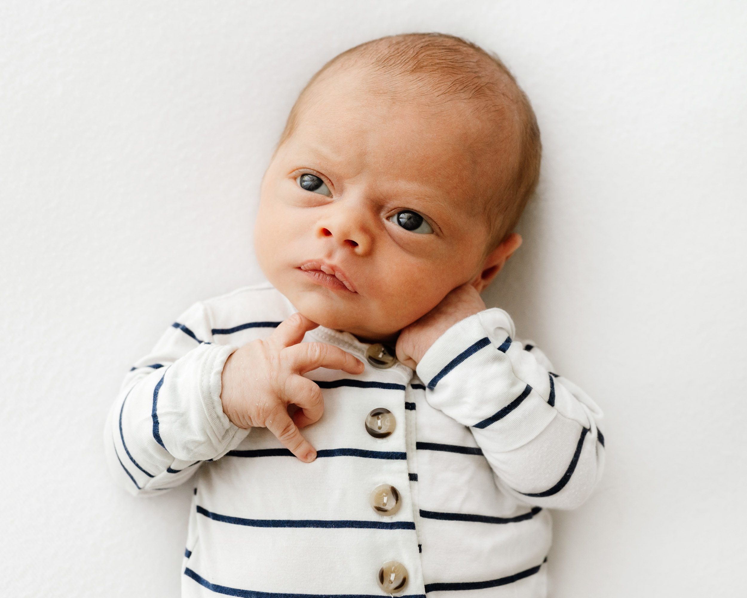a baby boy wearing a blue and white striped romper laying on a white backdrop and looking up toward the window light as he touches his chin with his finger during a newborn photo session