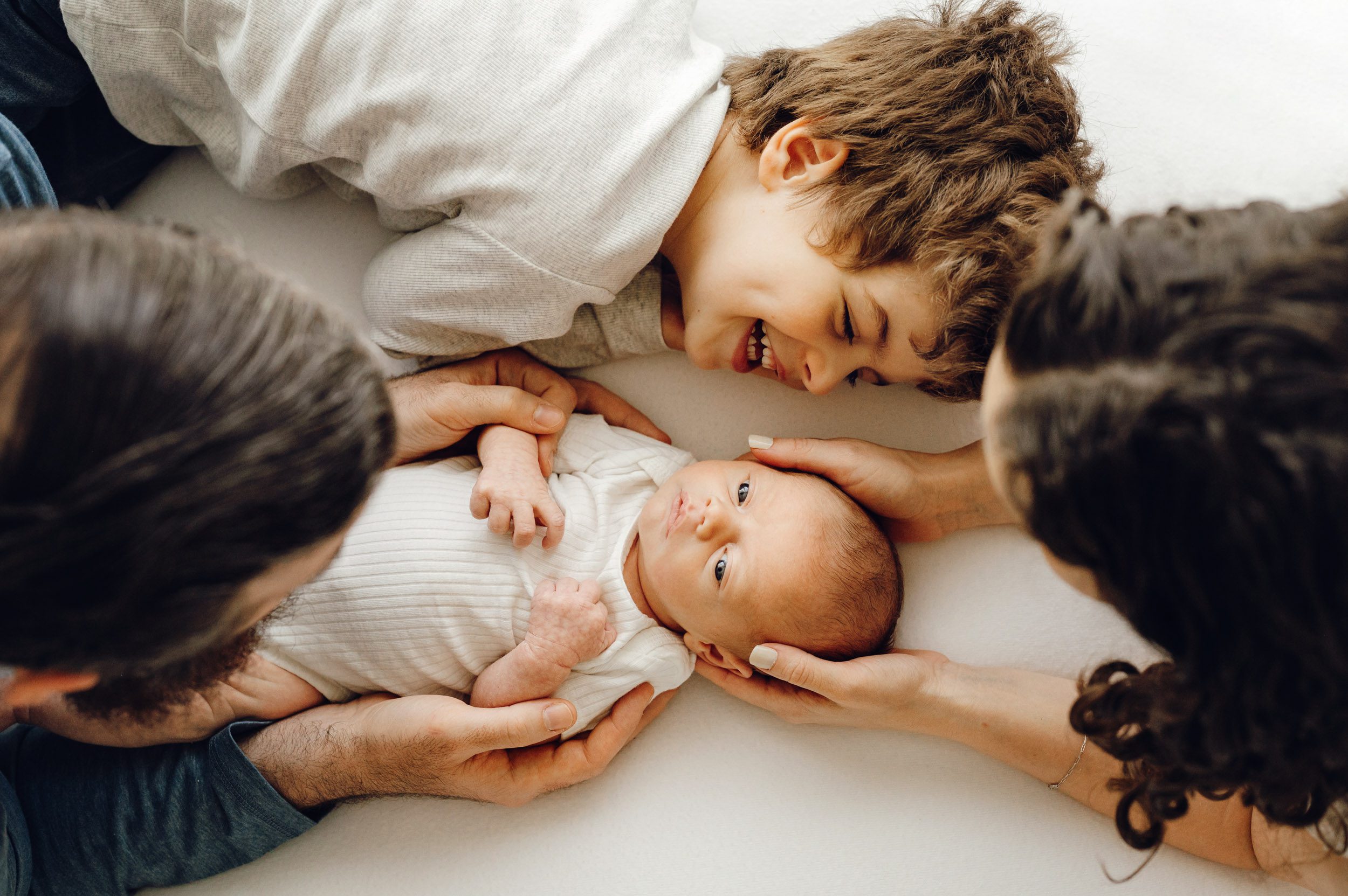 a picture taken from above of a baby boy laying on a white bean bag with his big brother laying right next to him and smiling at him as mom and dad look down at them during a newborn photoshoot