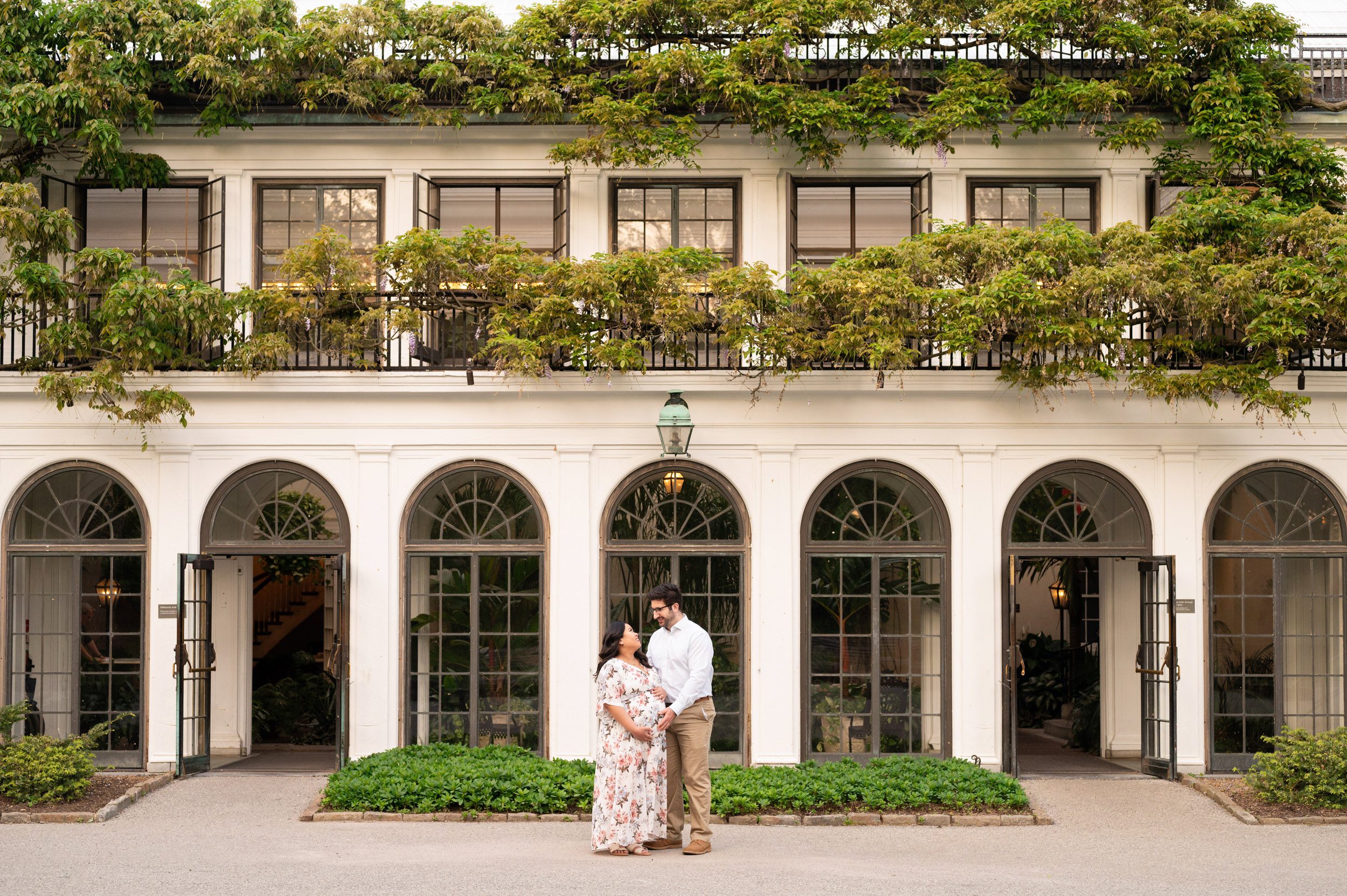an expecting couple standing in front of Dupont's House at Longwood Gardens with the big white arches and green wisteria vines growing across the building in the backdrop during a maternity photography session