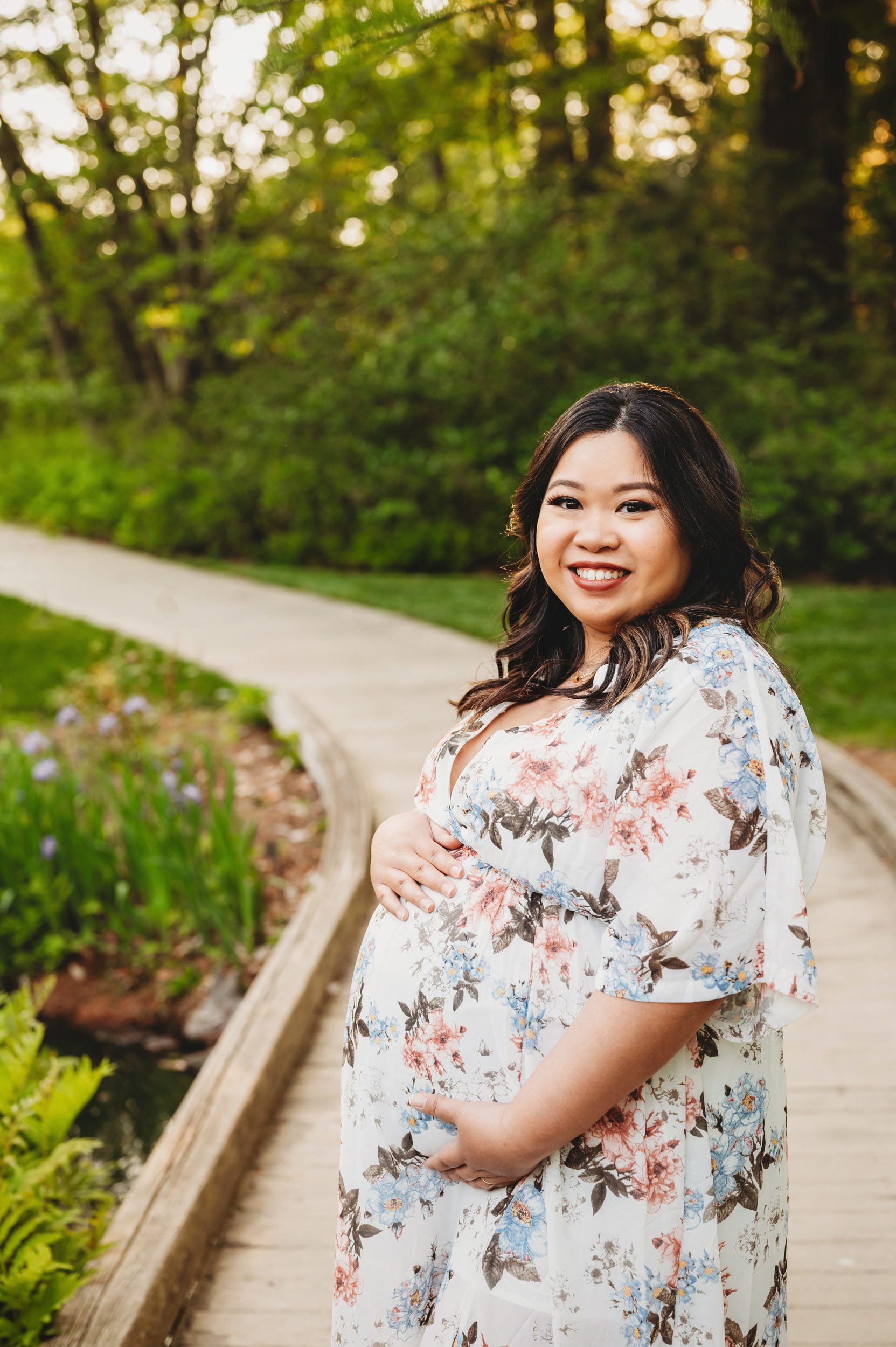 an expecting mother wearing a floral dress standing on a bridge and cradling her belly as she smiles at the camera during a Main Line maternity photo session