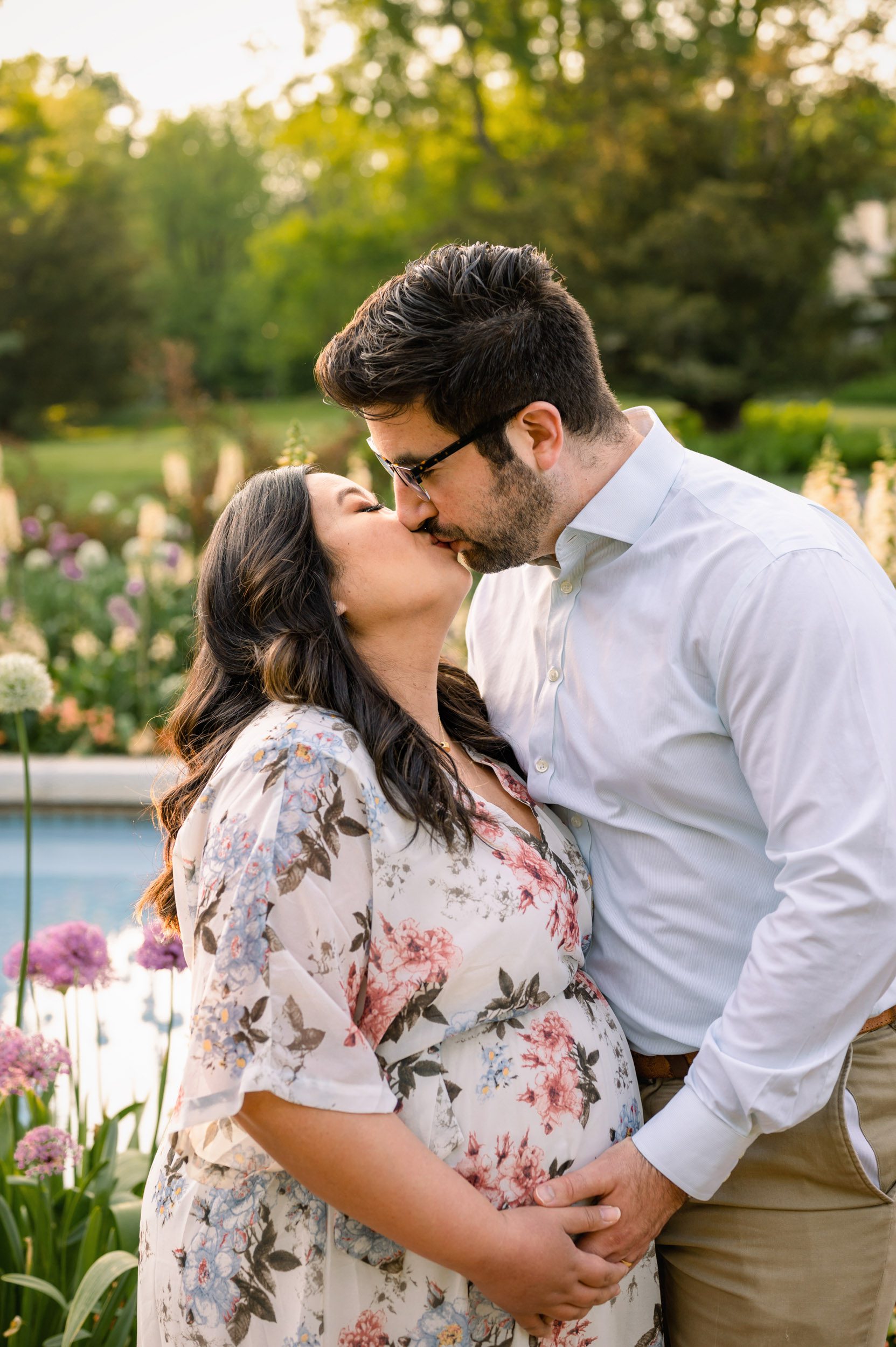 expecting parents standing in front of a fountain surrounded by flowers in bloom and kissing each other as they both touch mom's belly during a Main Line maternity session