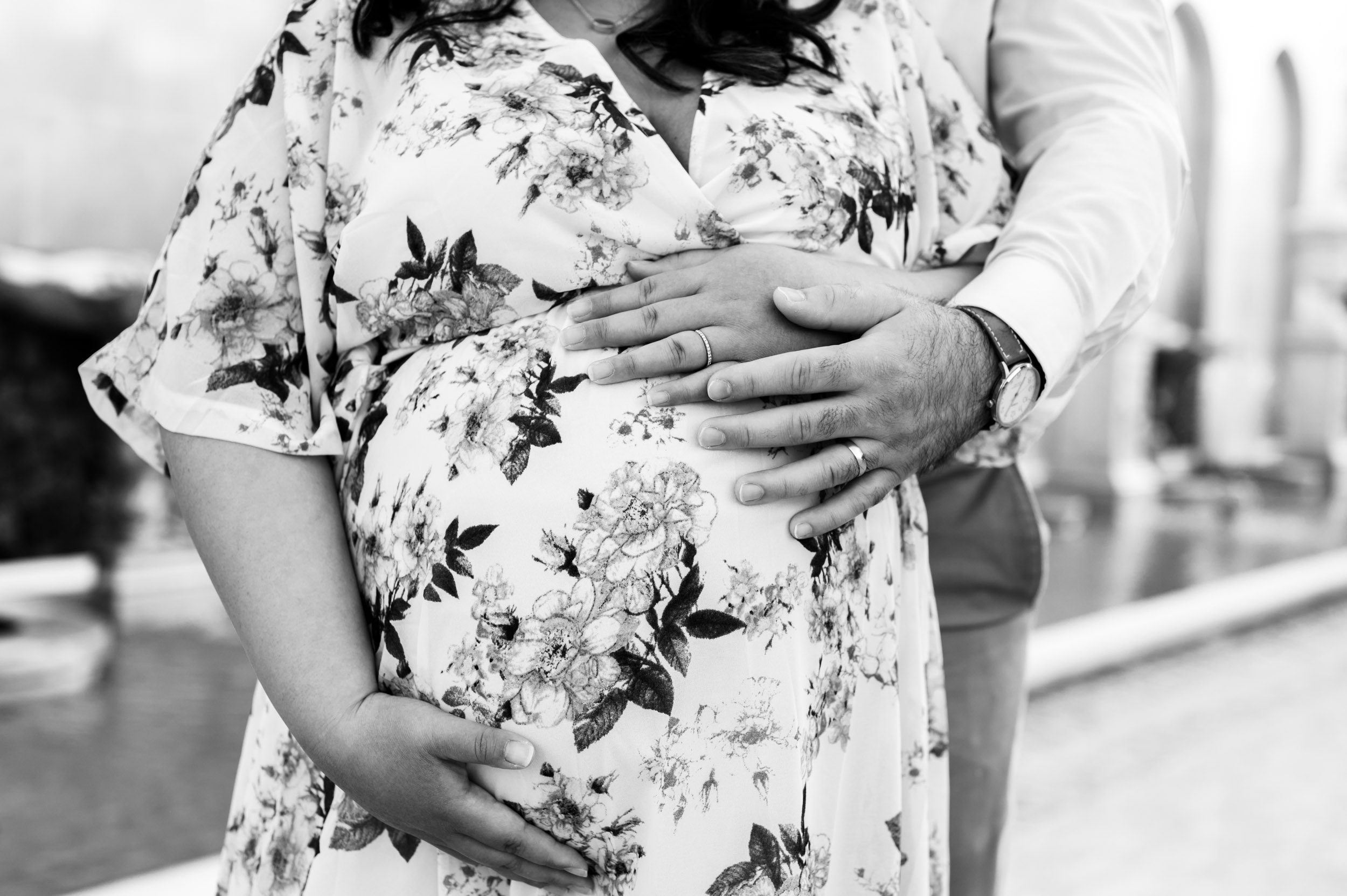 a black and white close up picture of an expecting mother wearing a floral print dress as her partner hugs her from behind and they both embrace her belly during a Main Line maternity photoshoot