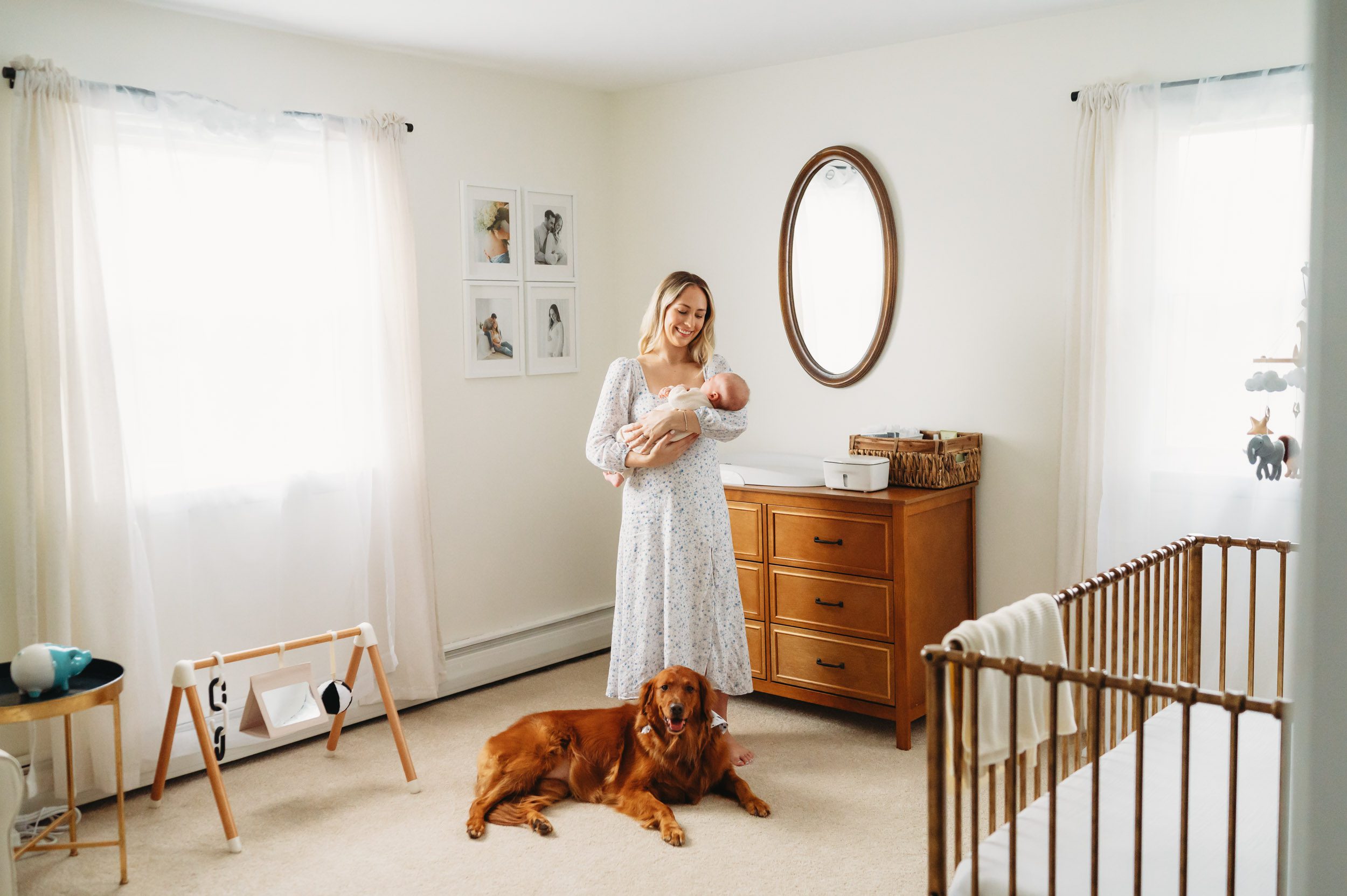a new mother standing in her son's nursery holding her baby boy in her arms and smiling down at him while the family dog sits at her feet and looks at the camera during an in home newborn photography session
