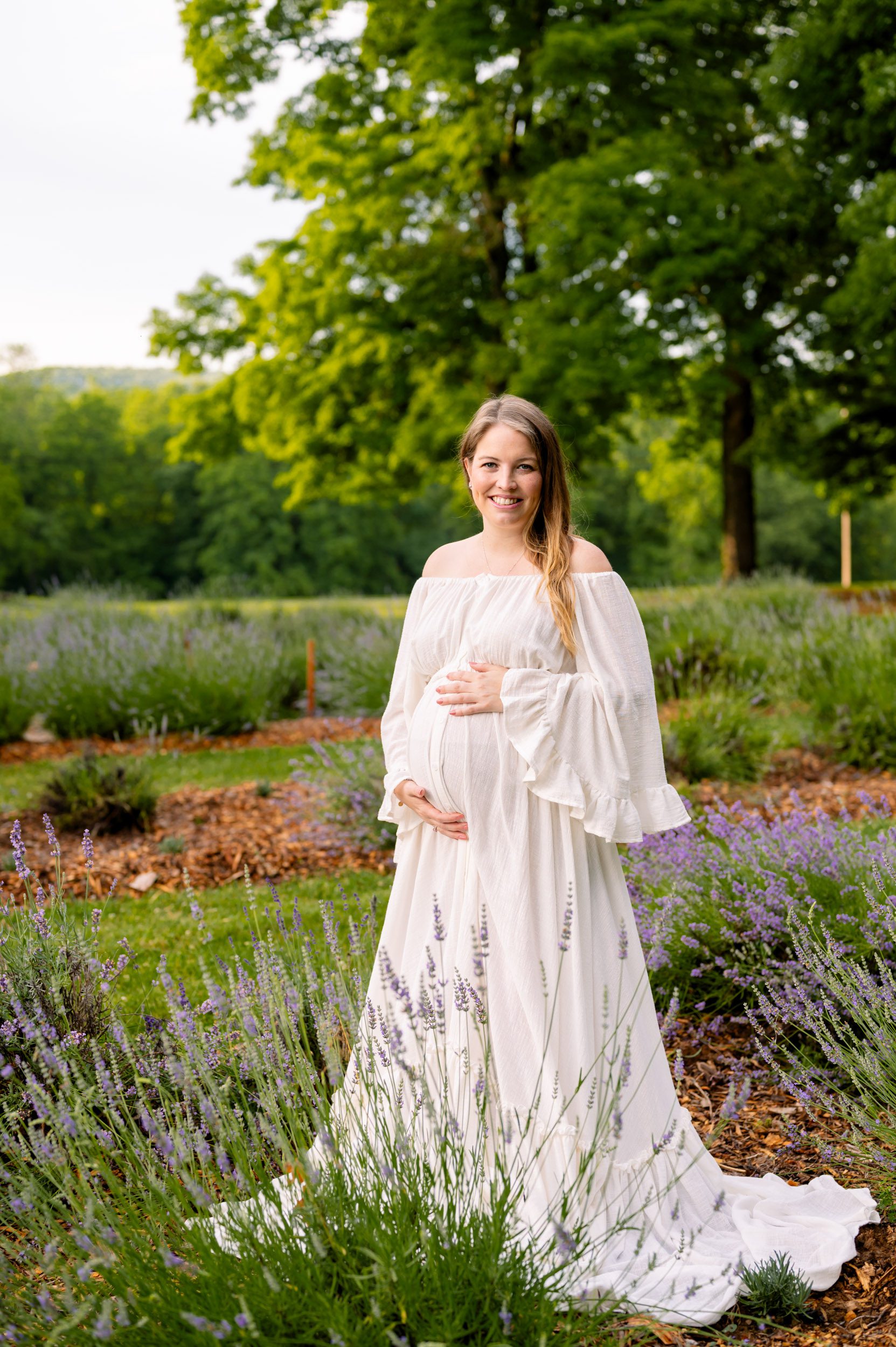 an expecting mother standing wearing a long, flowy white dress and standing in a lavender field and cradling her belly as she looks at the camera and smiles during a maternity photoshoot