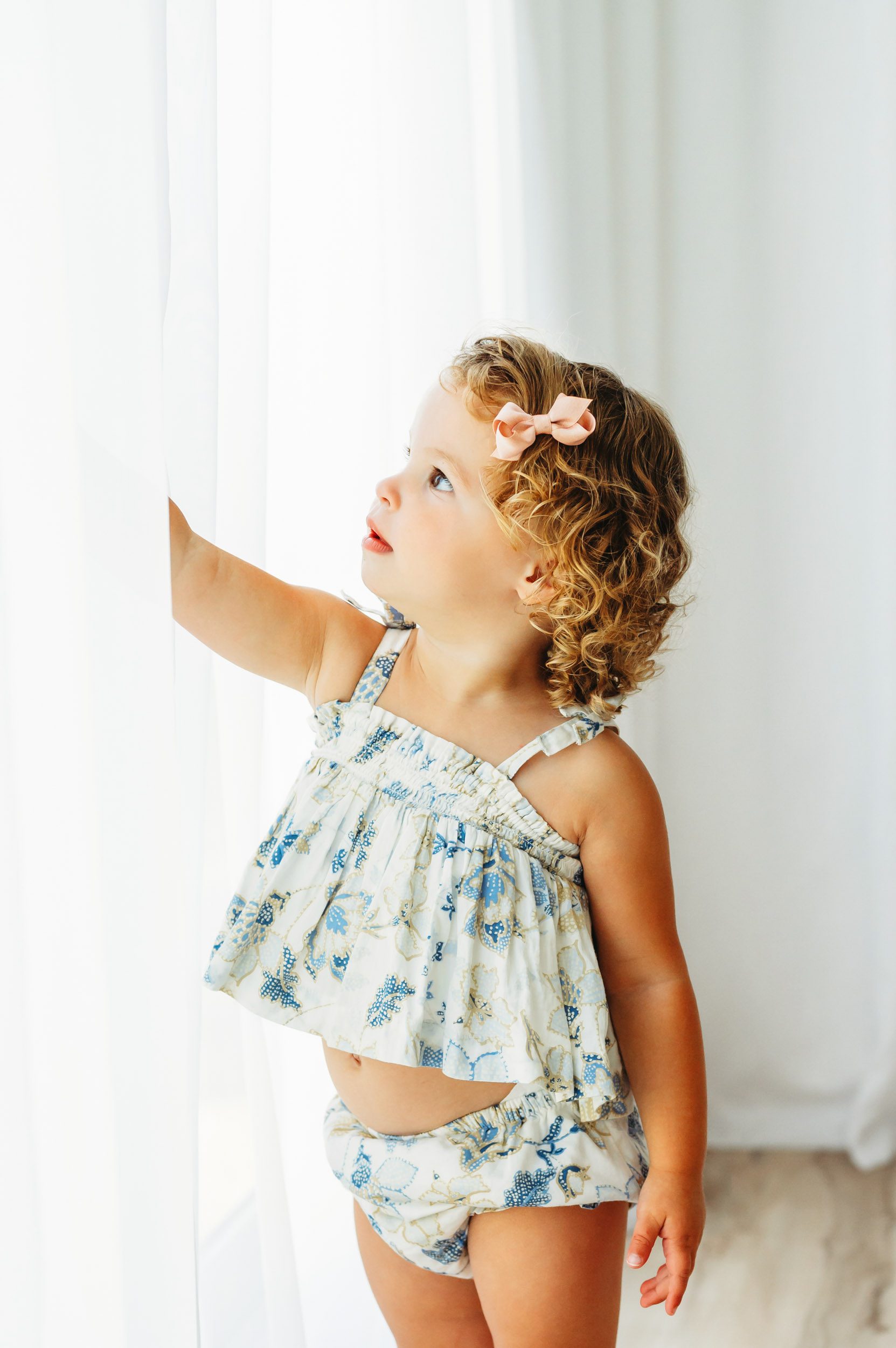 a toddler wearing a blue and gold floral outfit standing in front of a wall of windows and reaching up to touch the curtains during a 2nd birthday photoshoot