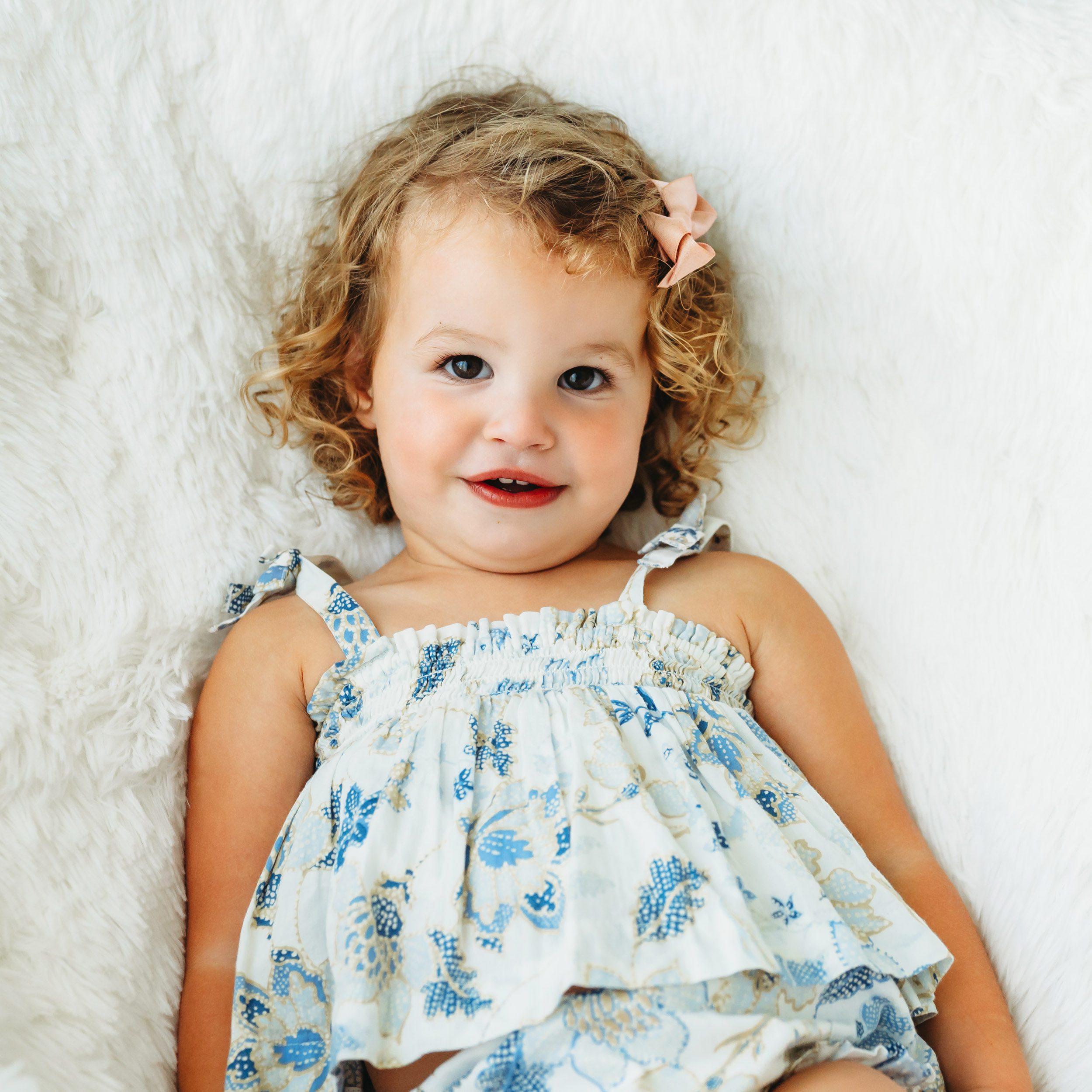 a young girl wearing a blue and gold floral top and a pink bow in her curly hair sitting in a white fluffy chair and smiling right at the camera during a 2nd birthday photoshoot