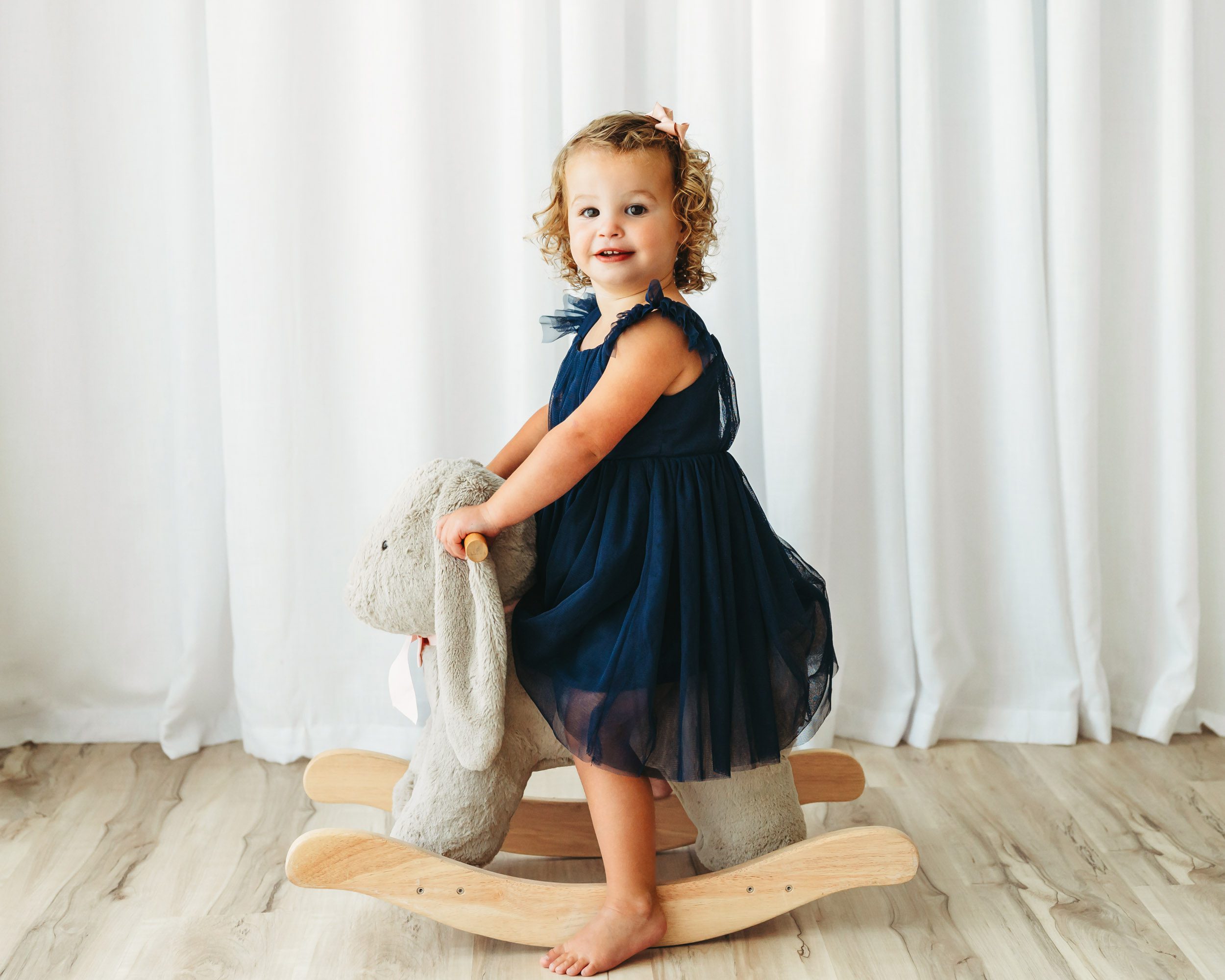 a little girl wearing a blue dress riding on a bunny rocker toy and smiling at the camera during a 2nd birthday photoshoot