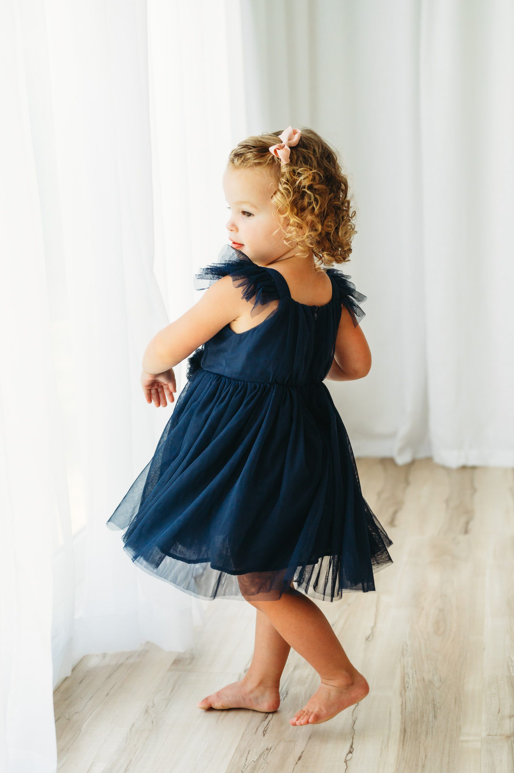 a toddler wearing a blue dress standing in front of a wall of windows and playing with the sheer curtains during a 2nd birthday photoshoot