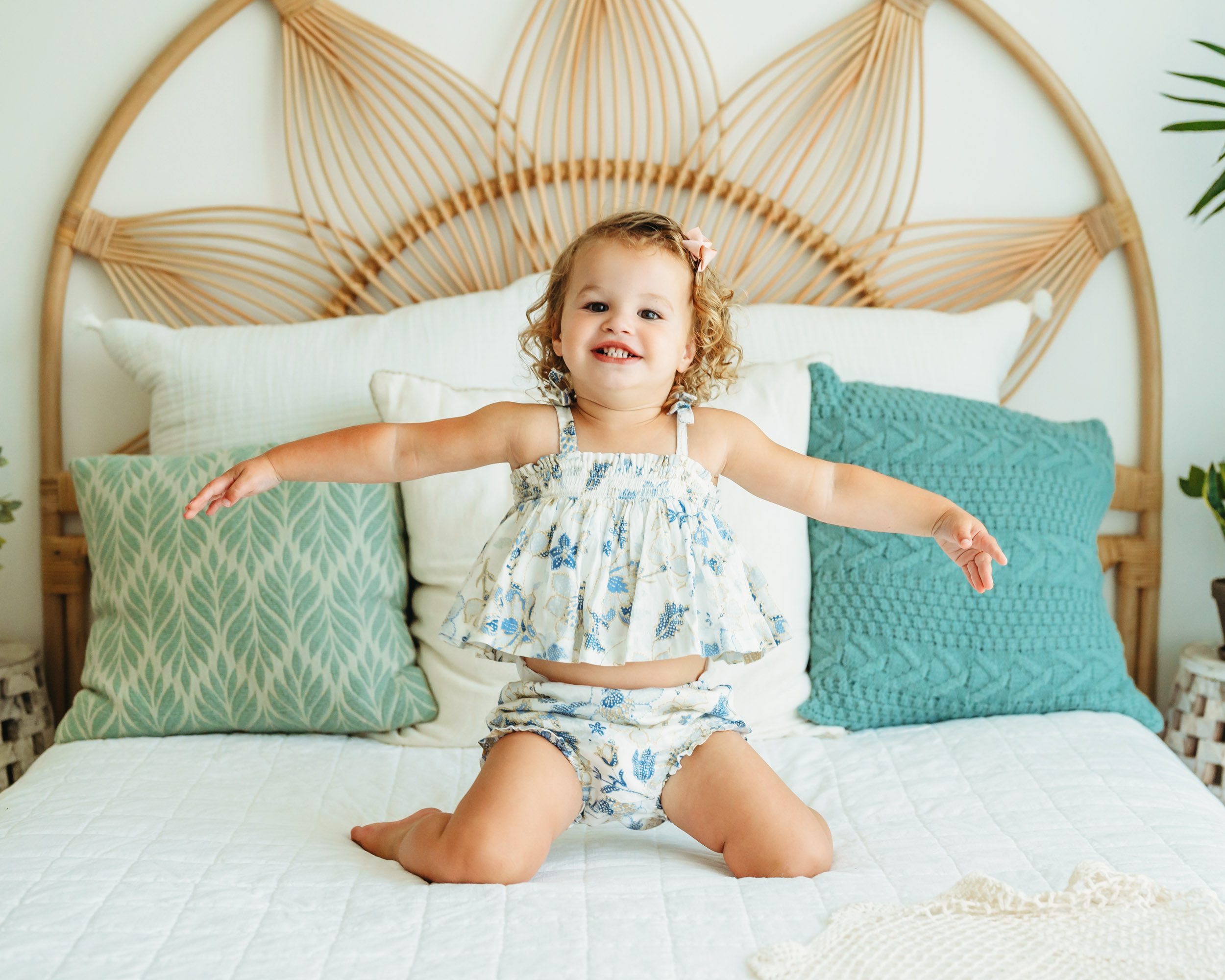 a little girl wearing a blue and gold floral outfit bouncing up and down on her knees on a bed during a toddler milestone photoshoot
