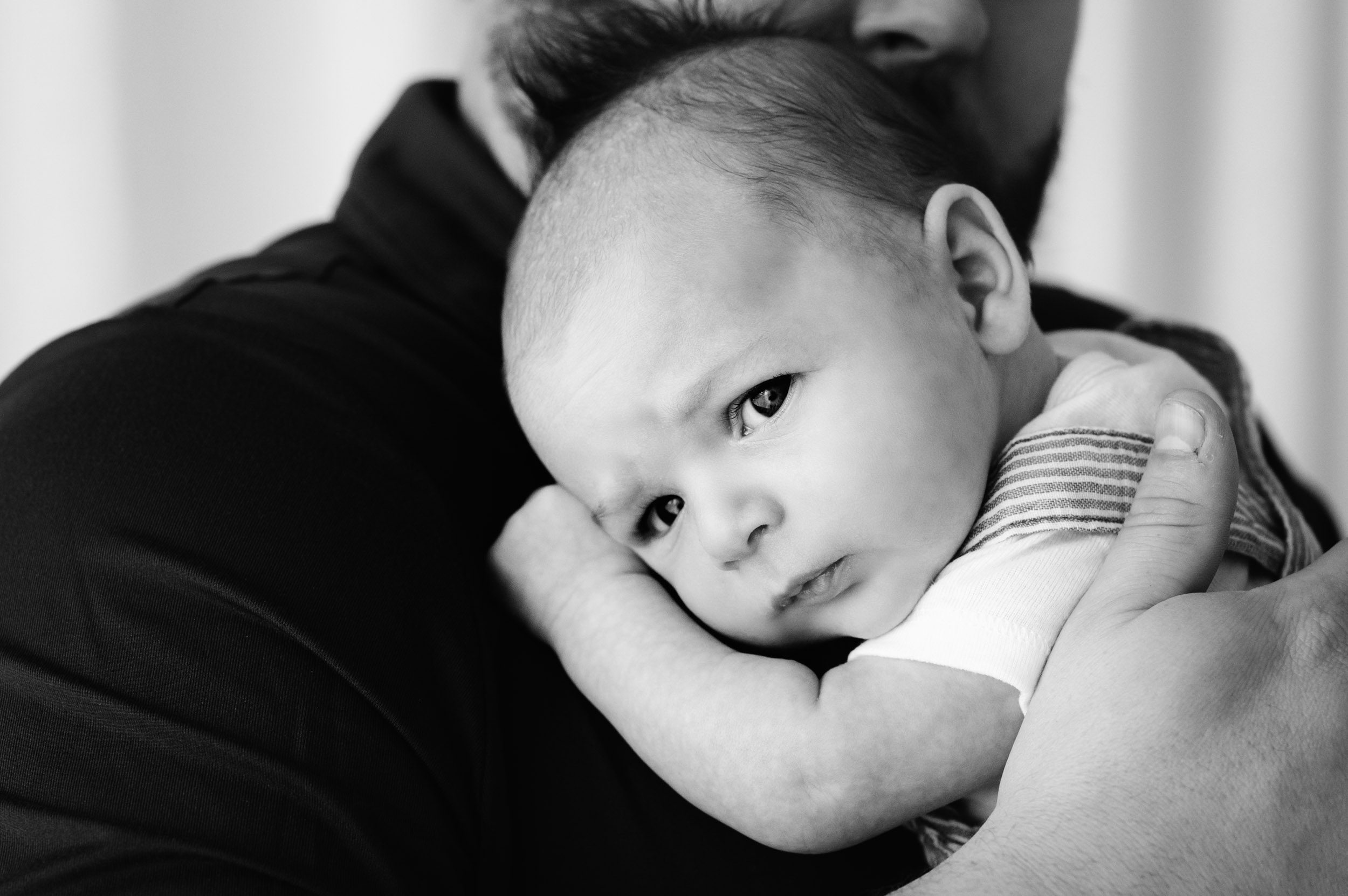 a black and white close up picture of a baby boy resting his head on his dad's shoulder and looking at the camera with a serious expression on his face during a newborn photoshoot