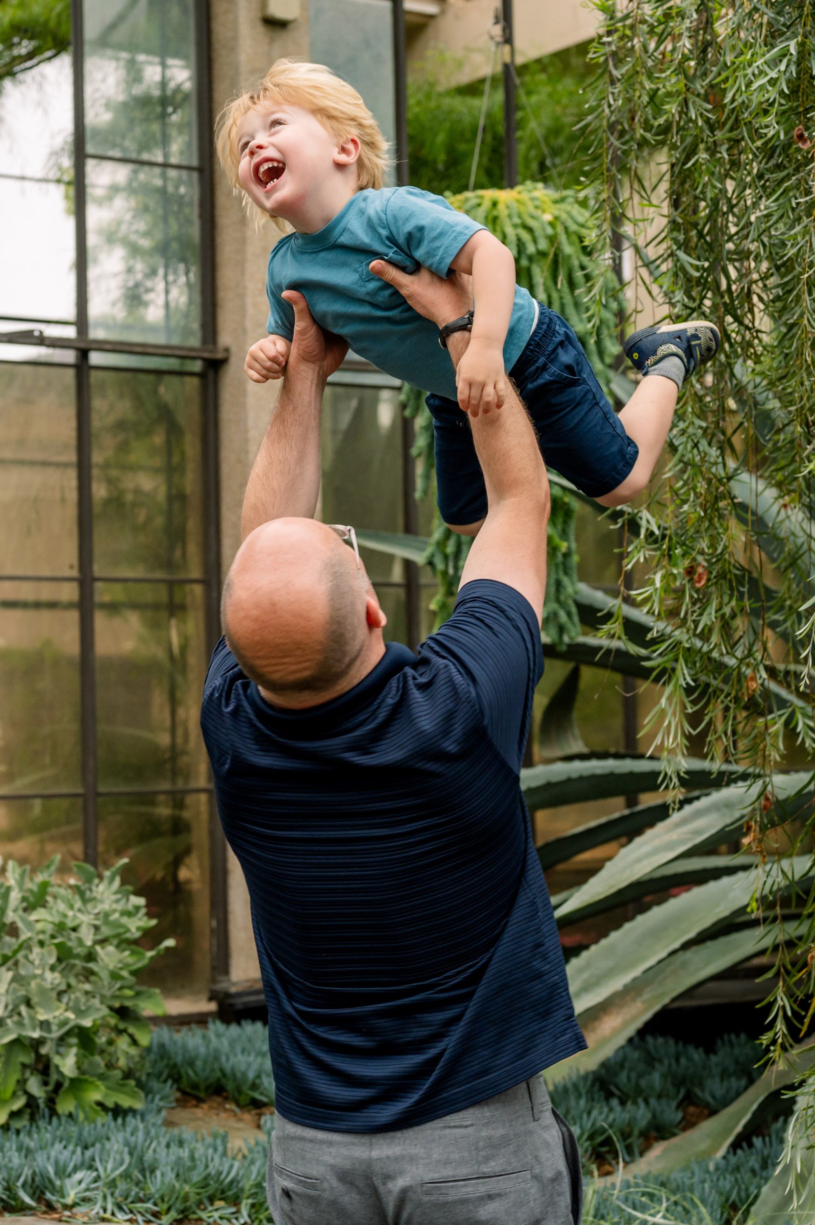 a dad standing in a greenhouse and lifting his young son high up in the air as he laughs during a Philadelphia family photoshoot
