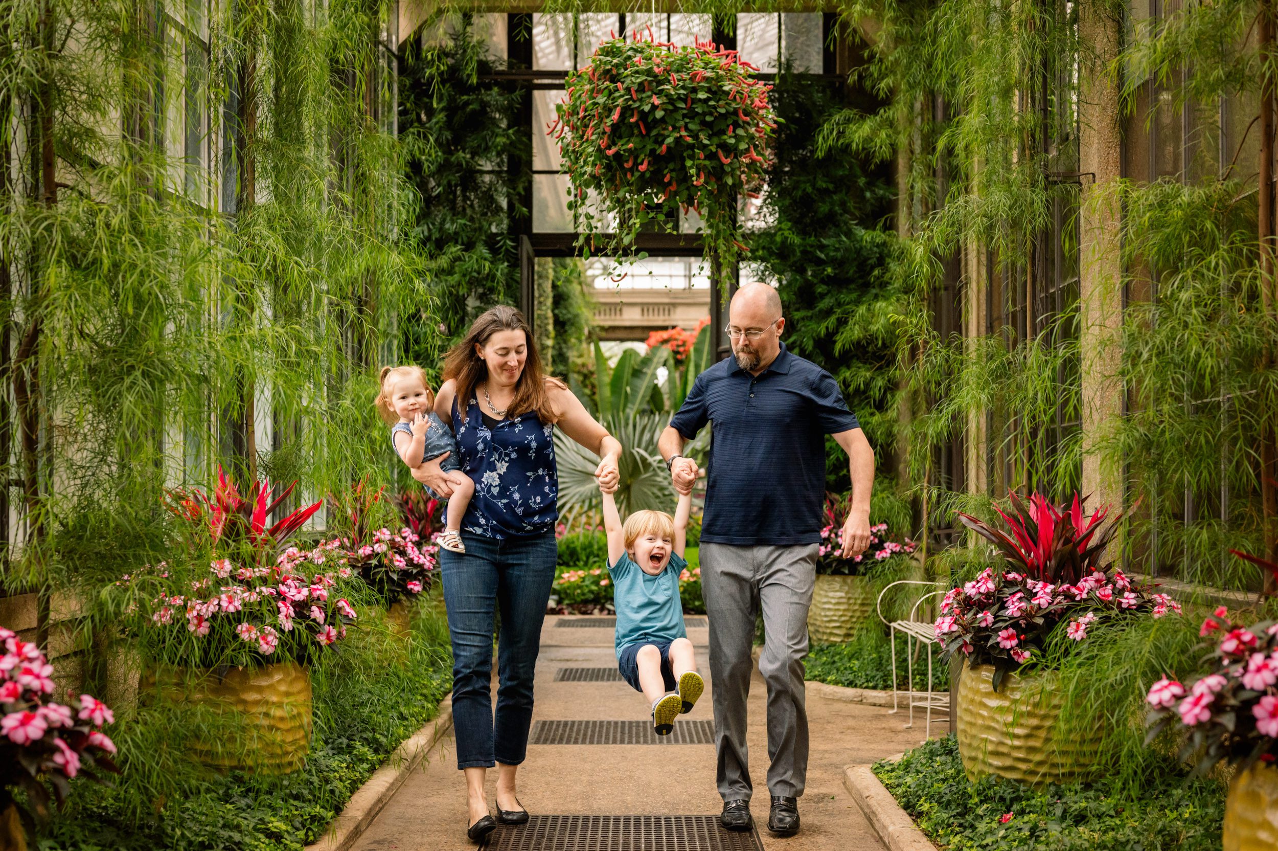 a family of four walking through a greenhouse surrounded by flowers and hanging vines on both sides as mom and dad swing their older son up in the air between them during a family photo session
